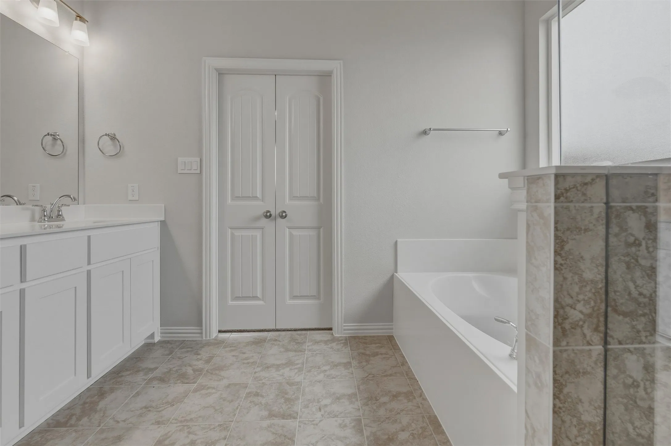 Dual sink vanity with white cabinetry, tile flooring, built-in bathtub, and double panel doors