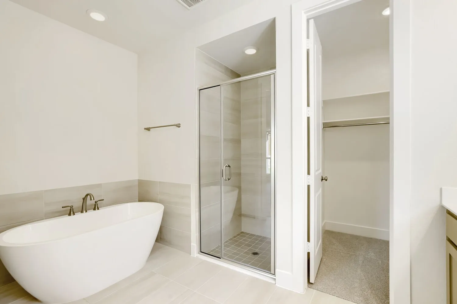 Freestanding soaking tub with a floor-mounted faucet, glass-enclosed shower with tiled walls and floor, recessed lighting, and a walk-in closet with carpeted flooring