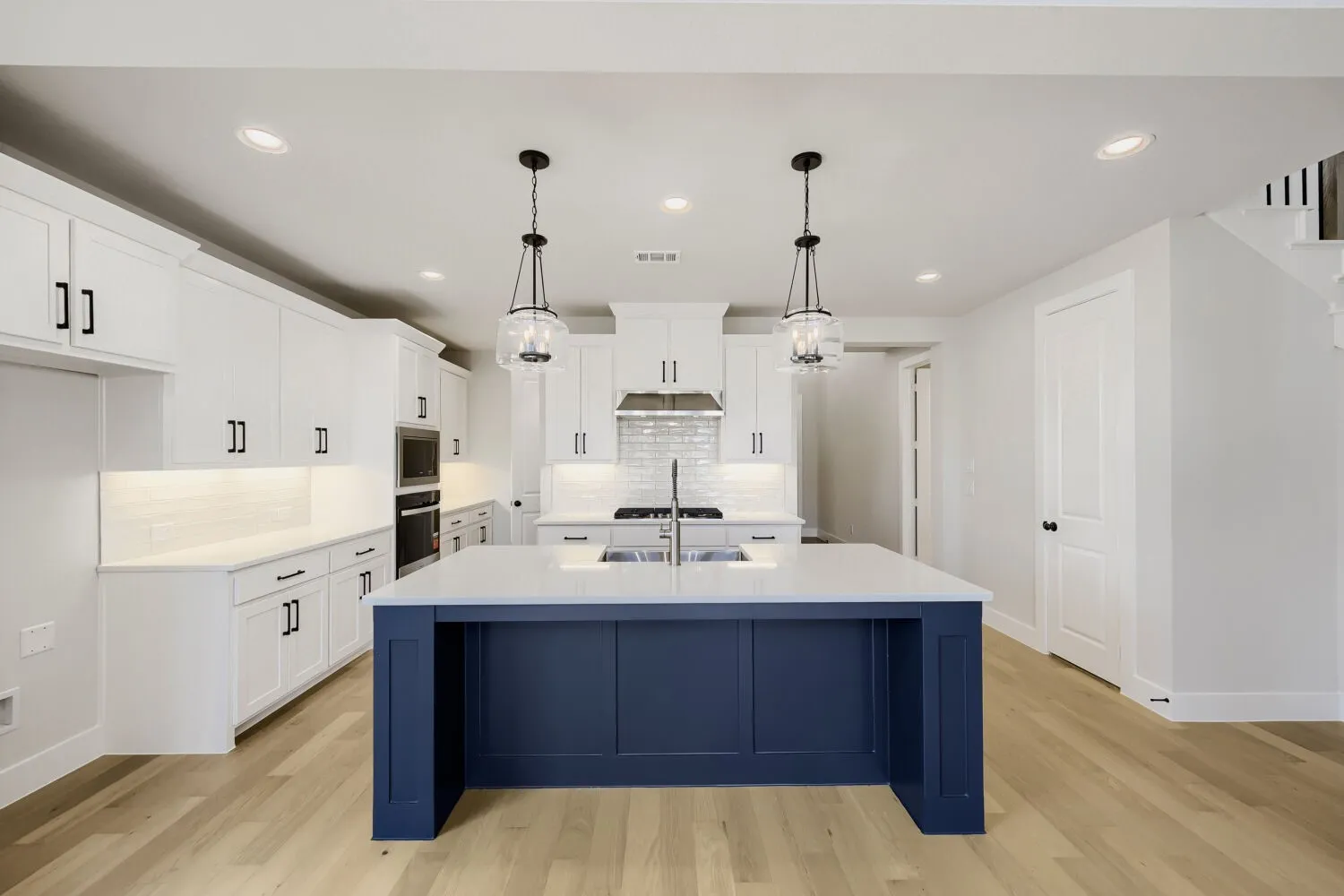 Chef's kitchen featuring white cabinetry with black hardware, a striking navy blue island with waterfall edge, stainless steel appliances including dual wall ovens, undermount sink with polished faucet, pendant lighting, recessed ceiling fixtures, and warm wood-finish flooring throughout the open layout