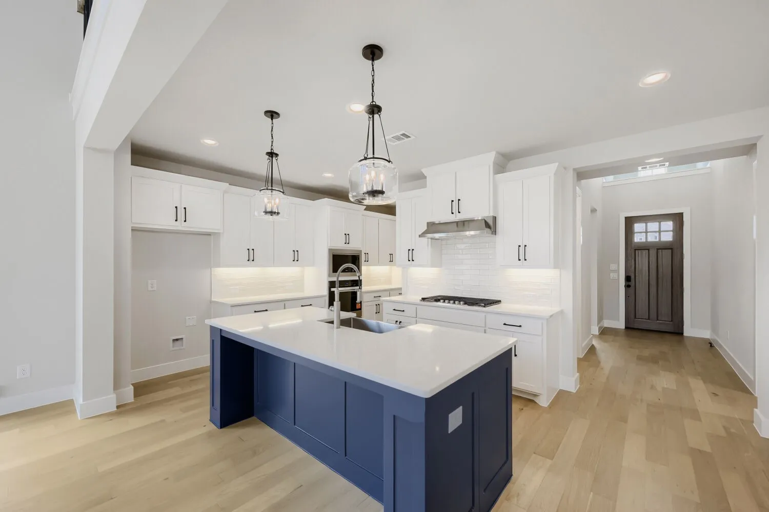 Contemporary kitchen featuring a custom navy blue island with white quartz countertops, white cabinetry with chrome hardware, stainless steel cooktop and range hood, pendant lighting with black metal fixtures, and wide-plank wood-finish flooring throughout the open layout