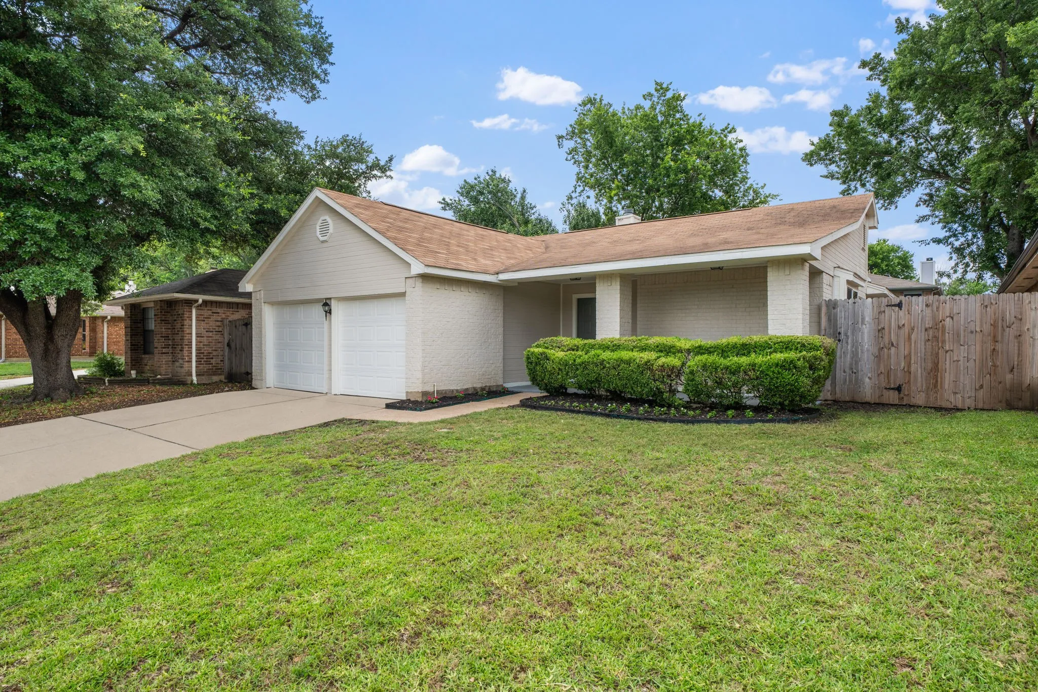Front exterior featuring light-colored brick siding, a two-car garage, a covered entry, a neatly trimmed shrub bed, and a wood privacy fence