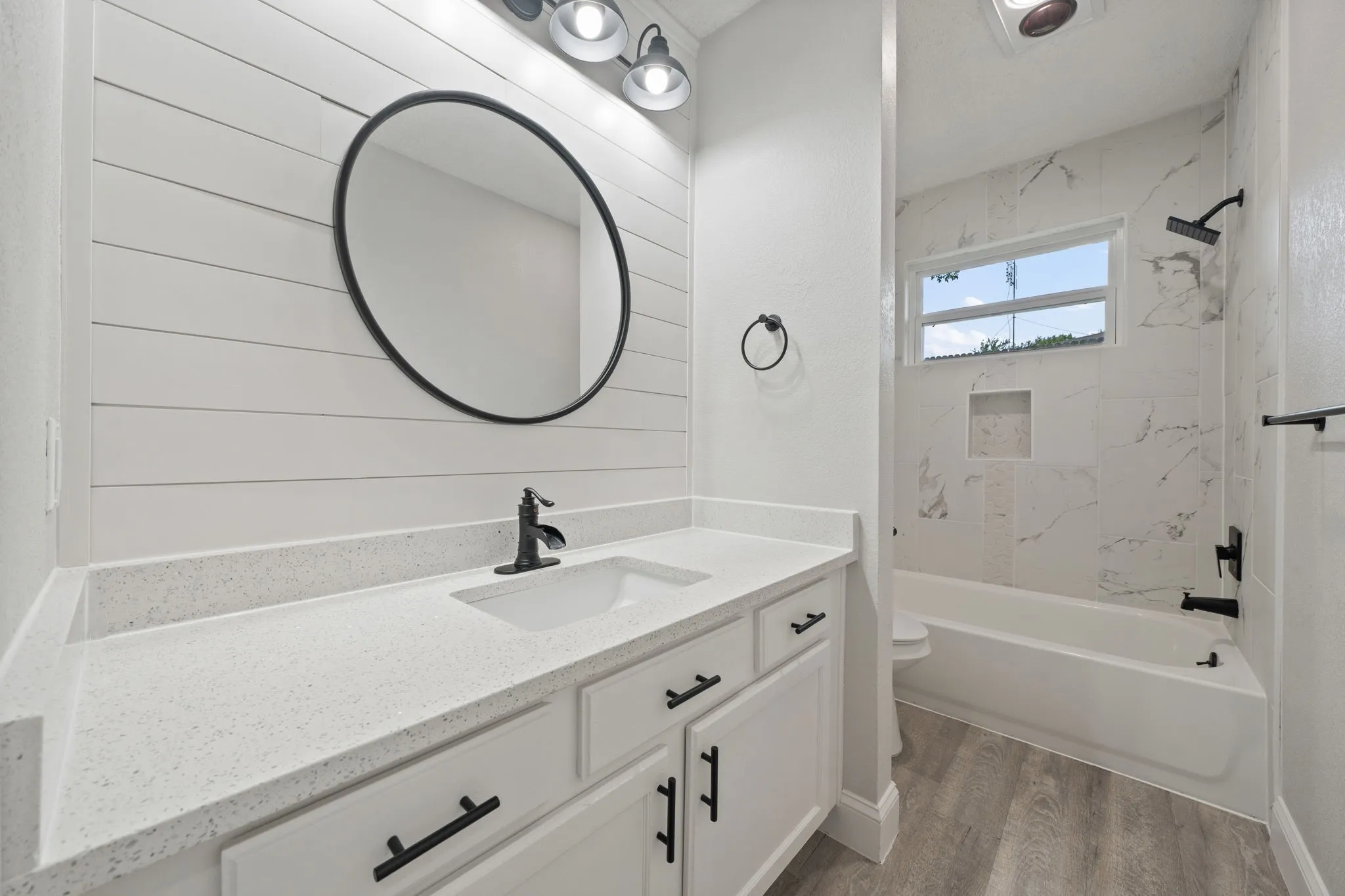 Bathroom featuring a white shiplap accent wall, white vanity with a light-toned countertop, black hardware and fixtures, a built-in tub with marble-look tile surround, and wood-finish flooring