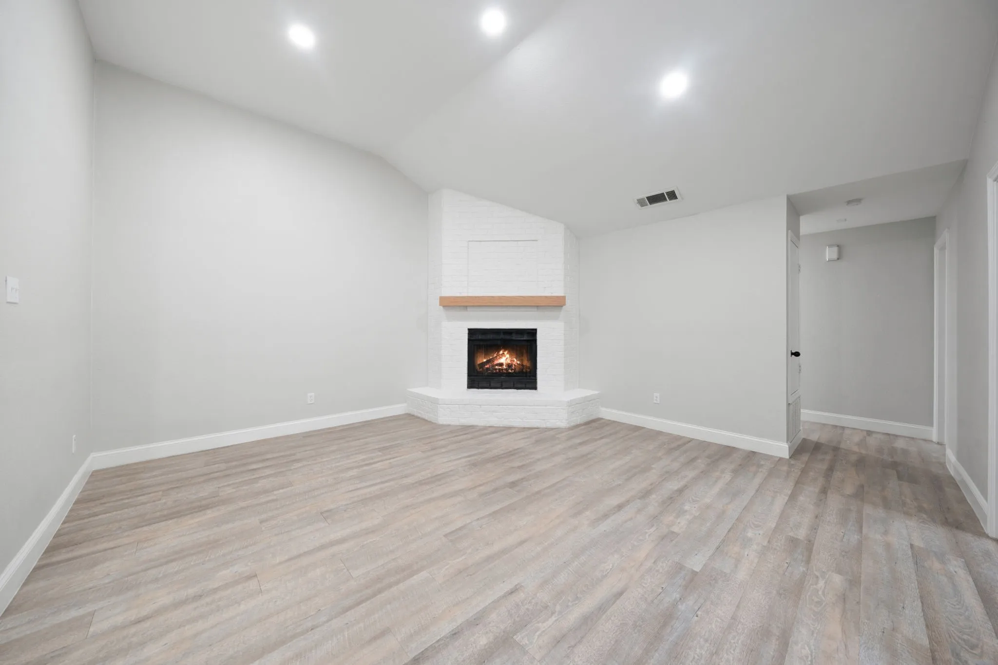 Spacious room featuring light wood-finish flooring, a white brick fireplace with a wood mantle, recessed lighting, and white baseboards