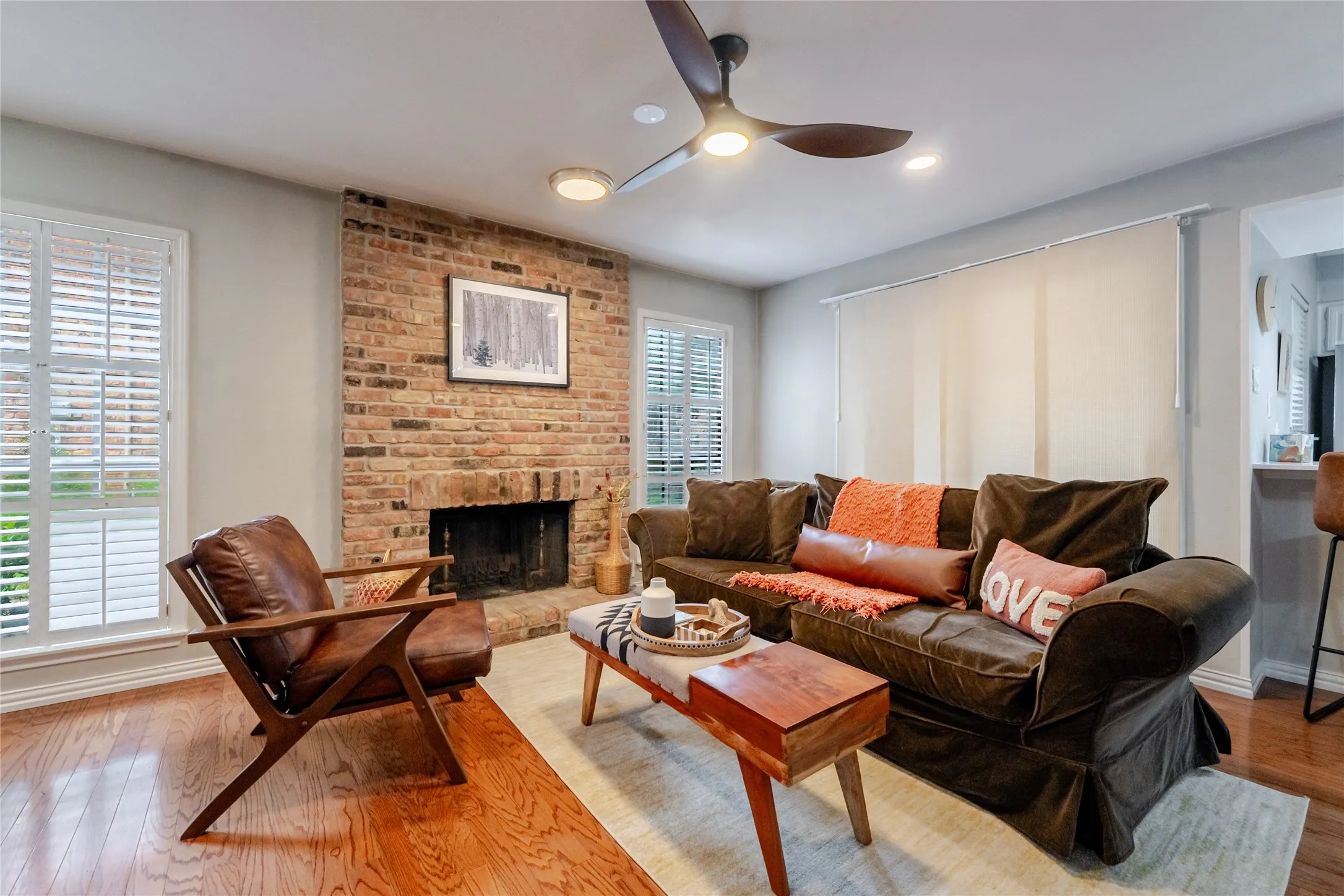 Living area featuring a brick fireplace, hardwood flooring, and white plantation shutters on dual windows