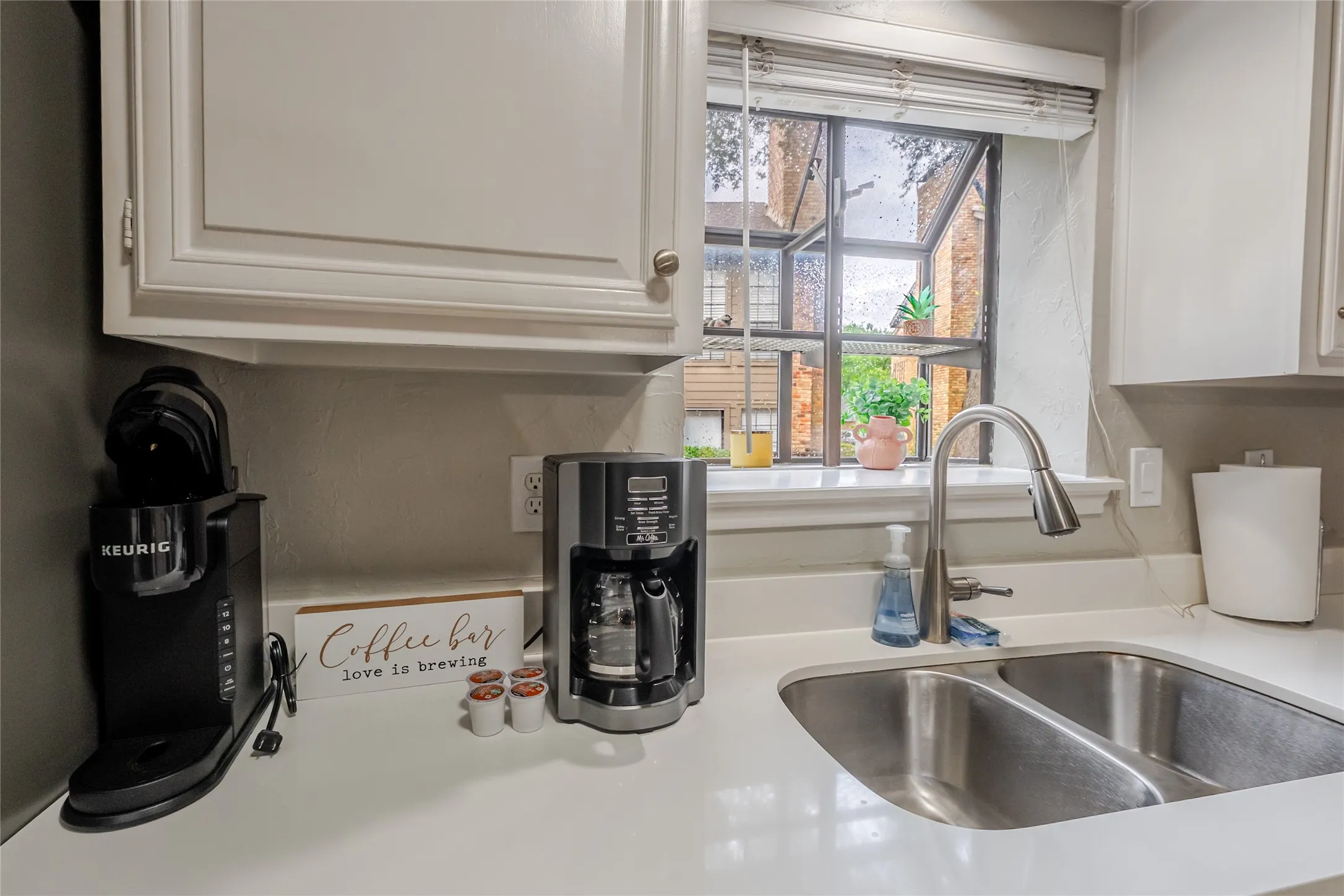 Kitchen window featuring a dual basin stainless steel sink, gooseneck faucet, light-toned countertops, and upper cabinetry with silver-tone hardware