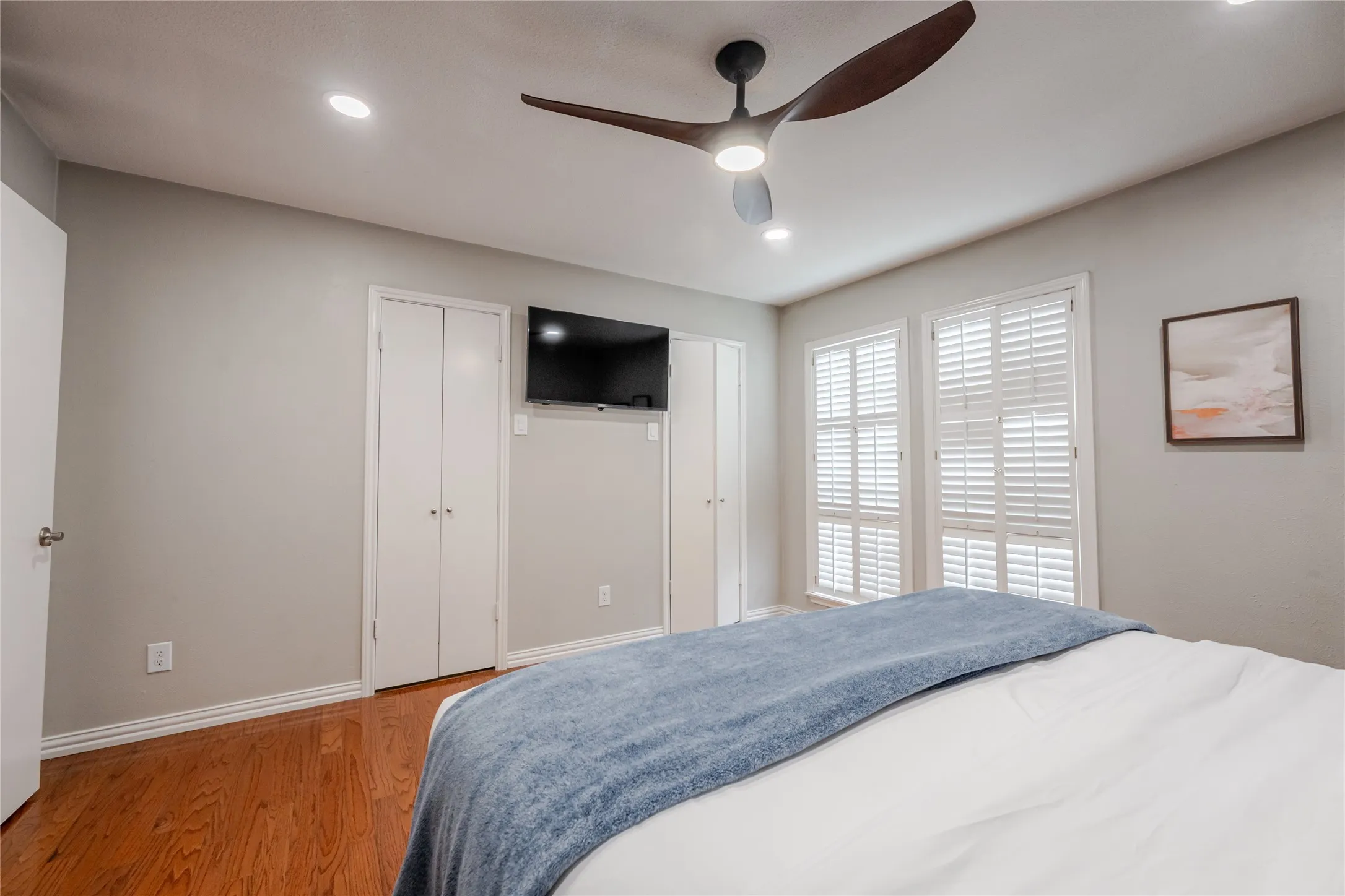 Bedroom featuring wood-finish flooring, two sets of white plantation shutters, and a modern ceiling fan with integrated lighting