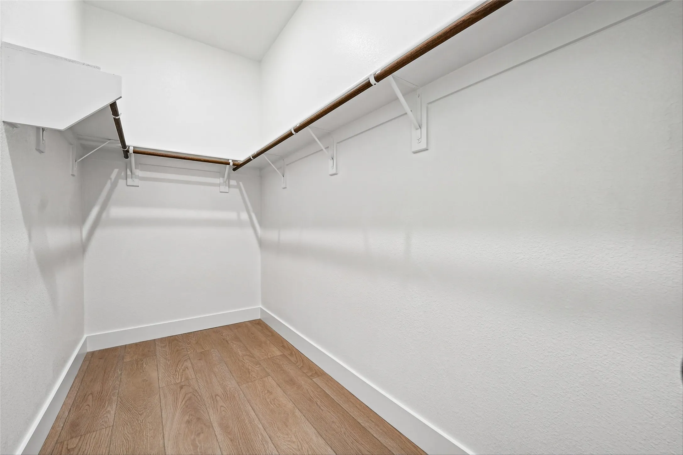 Walk-in closet featuring wood-finish flooring and L-shaped hanging rods with shelf above