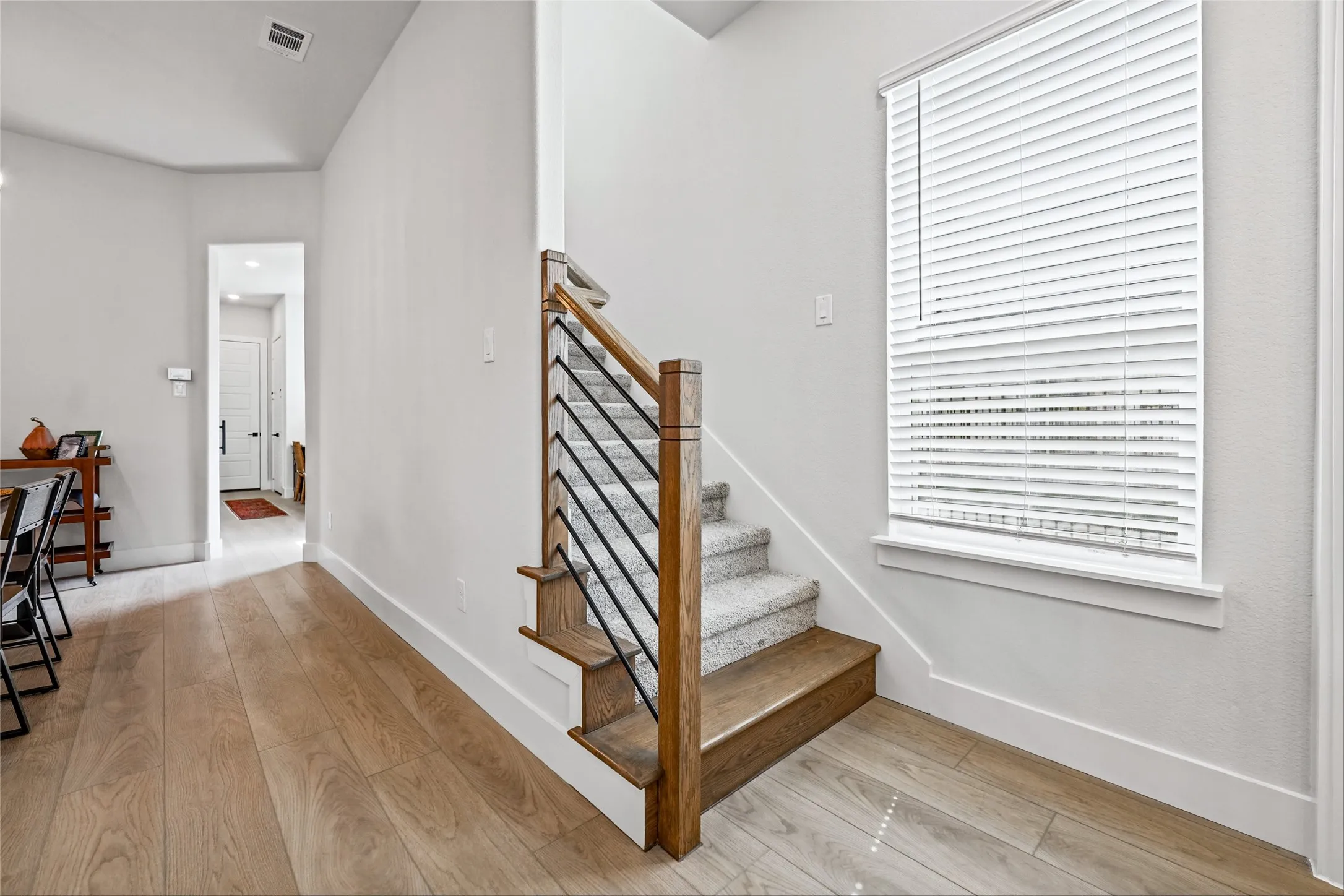 Wood-finish flooring, staircase with wood newel posts, black metal balusters, and carpeted treads, and a window with blinds