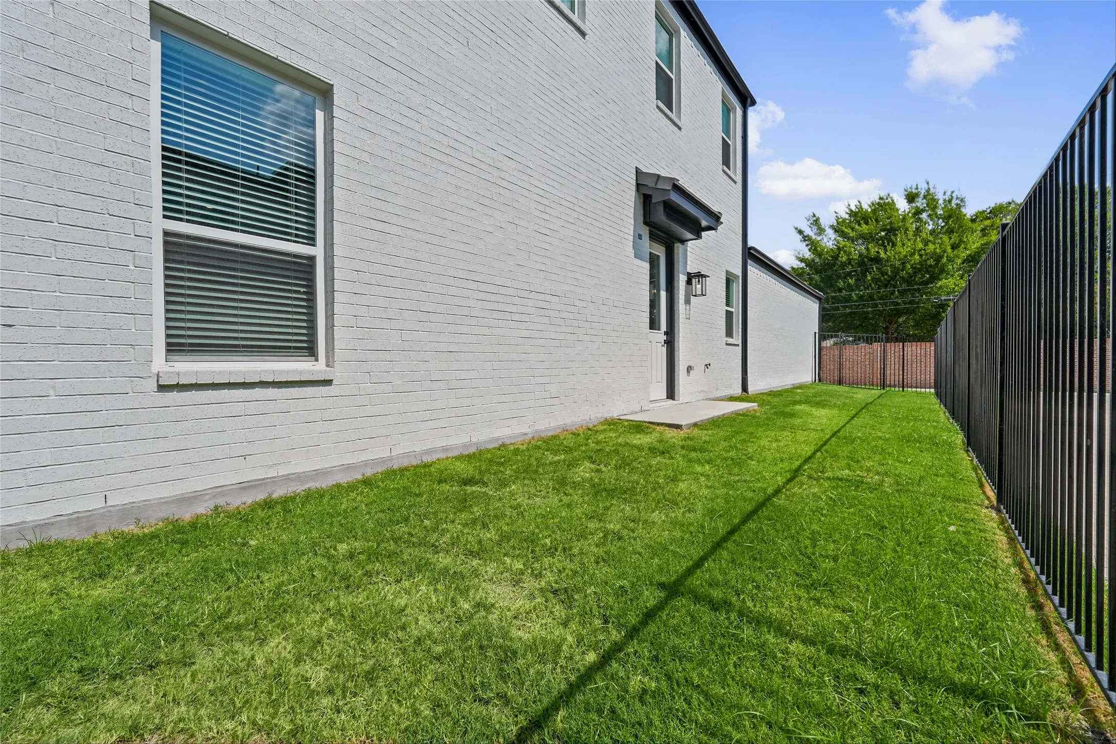 Painted brick exterior, side yard with green lawn, black metal fencing, exterior door with awning, and wall-mounted light fixture
