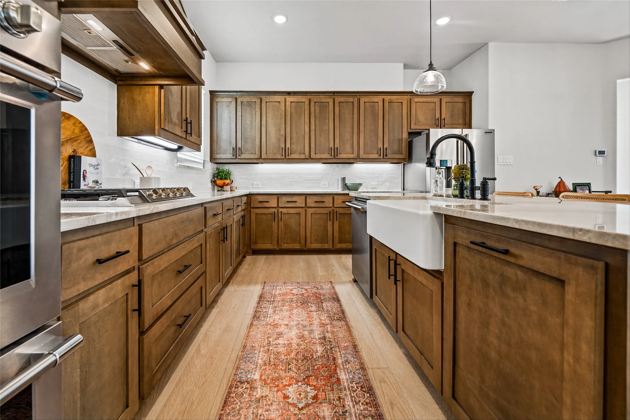 Gourmet kitchen featuring extensive wood cabinetry, light-toned countertops, a farmhouse sink, stainless steel appliances, and wood-finish flooring