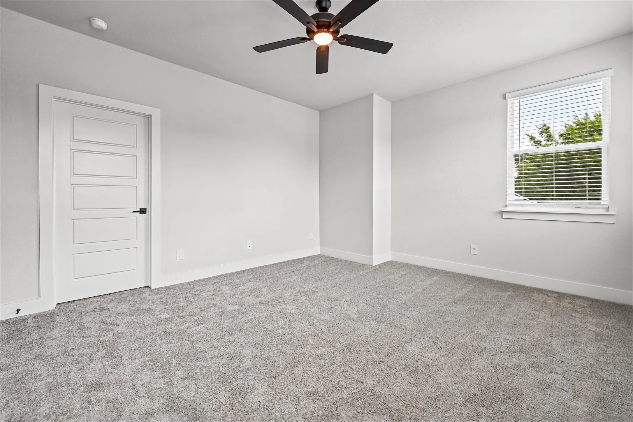 Neutral gray carpeting, white painted walls, a multi-panel door with black hardware, a ceiling fan with integrated lighting, and a window with blinds