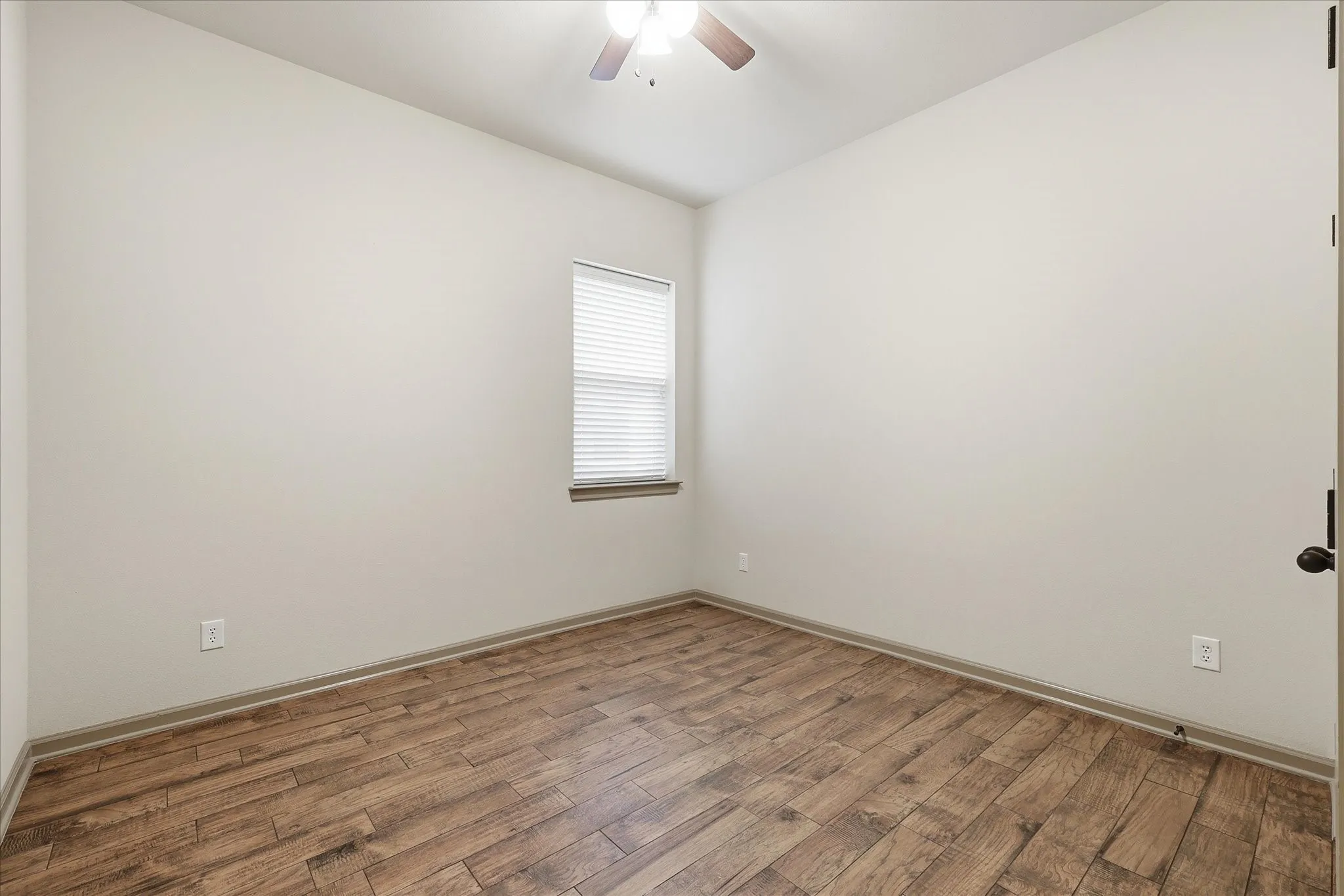 Downstairs secondary bedroom featuring wood-finish tile flooring, a window with blinds, a ceiling fan, light-colored walls, and a dark-toned door