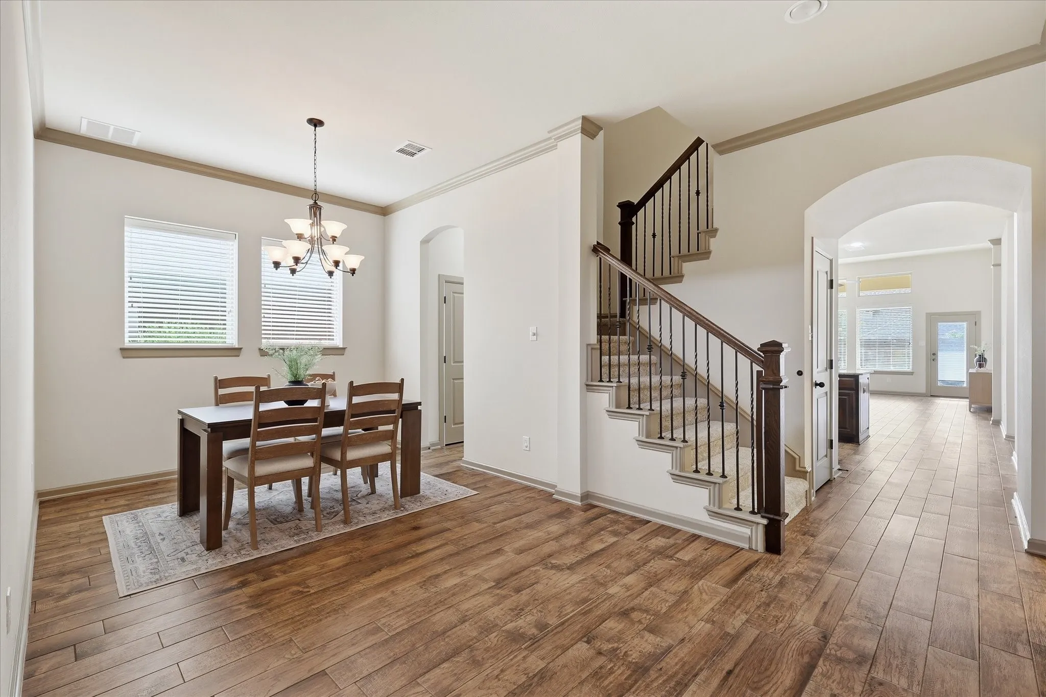 Spacious interior featuring wood-finish tile flooring, a chandelier, a staircase with dark wood banisters and metal spindles, and an arched doorway leading to the kitchen and living room