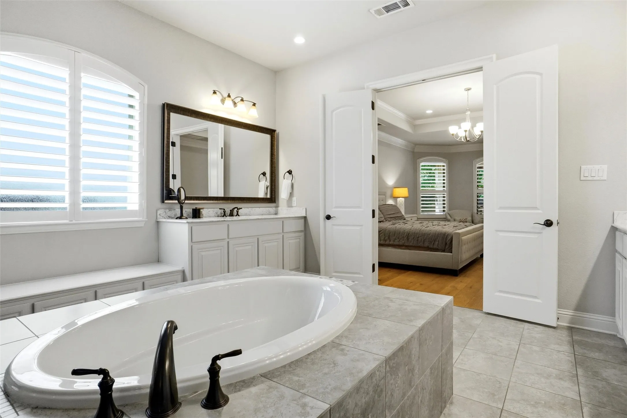 Bathroom featuring an oval drop-in tub with dark bronze fixtures, a double vanity with stone countertop and framed mirror, and an arched window with plantation shutters