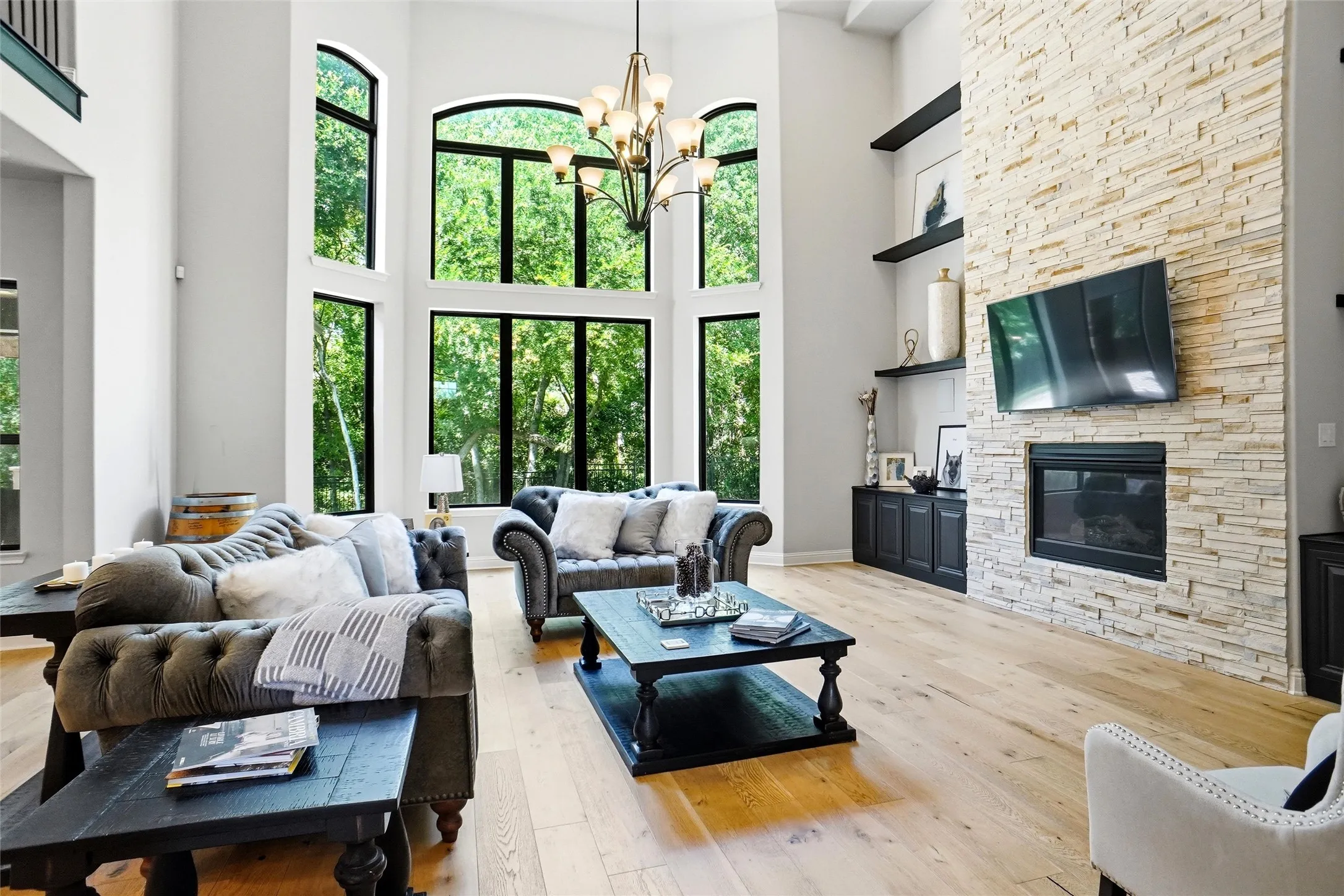 Double-height living space featuring expansive arched and rectangular windows, light wood-finish flooring, a stone-clad fireplace, built-in cabinetry, and integrated shelving