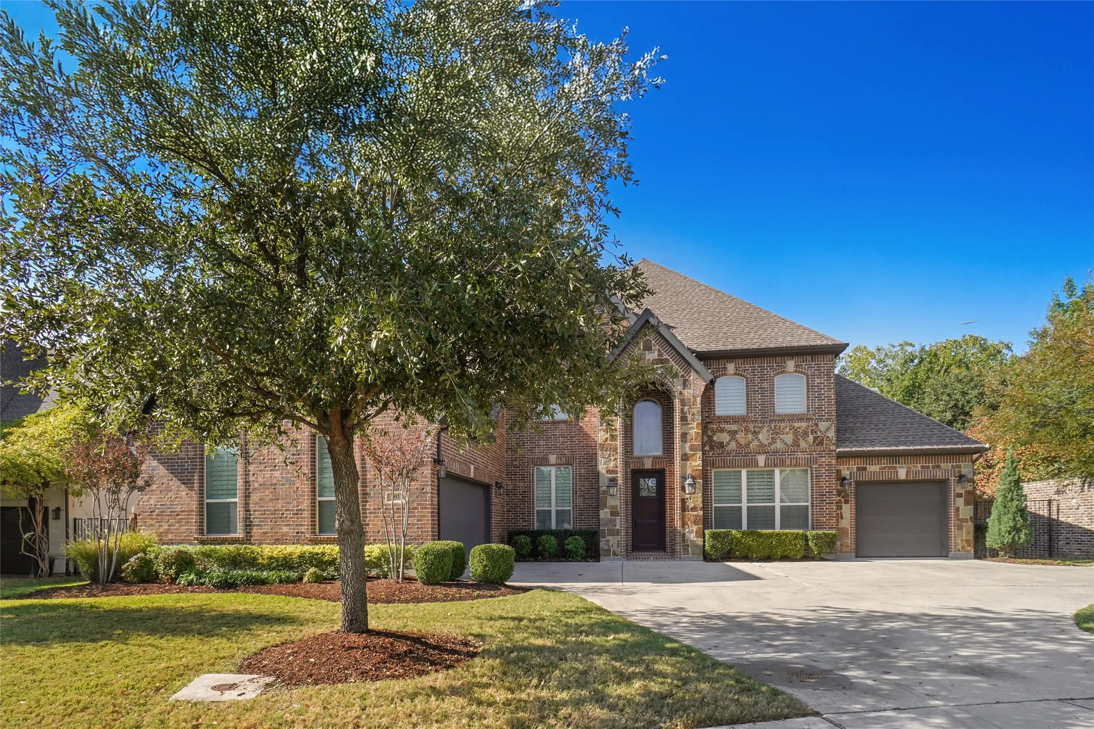 Brick and stone facade residence featuring a two-car garage, mature landscaping, and an arched entry
