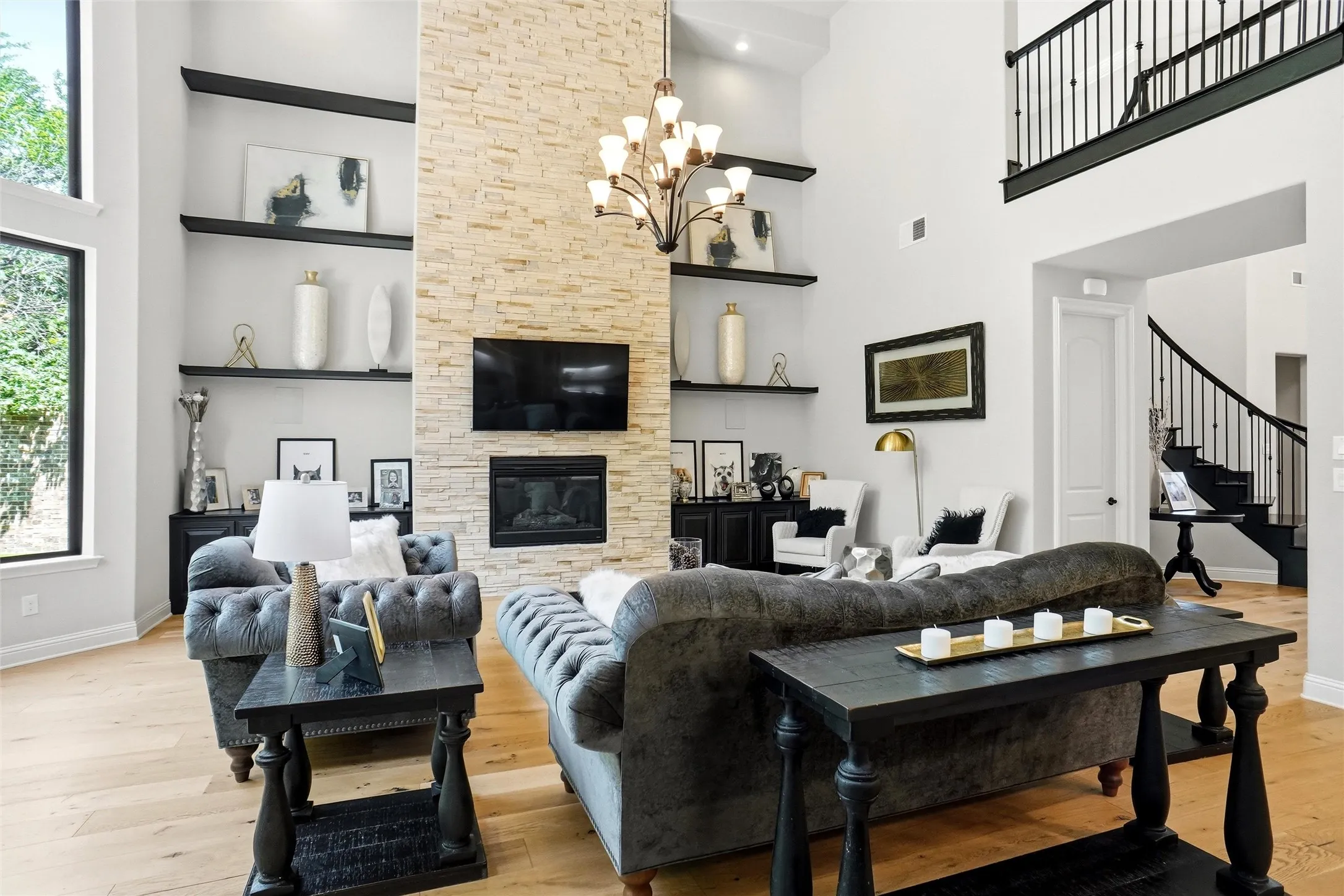 Two-story living area featuring a stone-clad fireplace with an integrated television, expansive windows, light wood-finish flooring, and a second-story balcony with metal railings