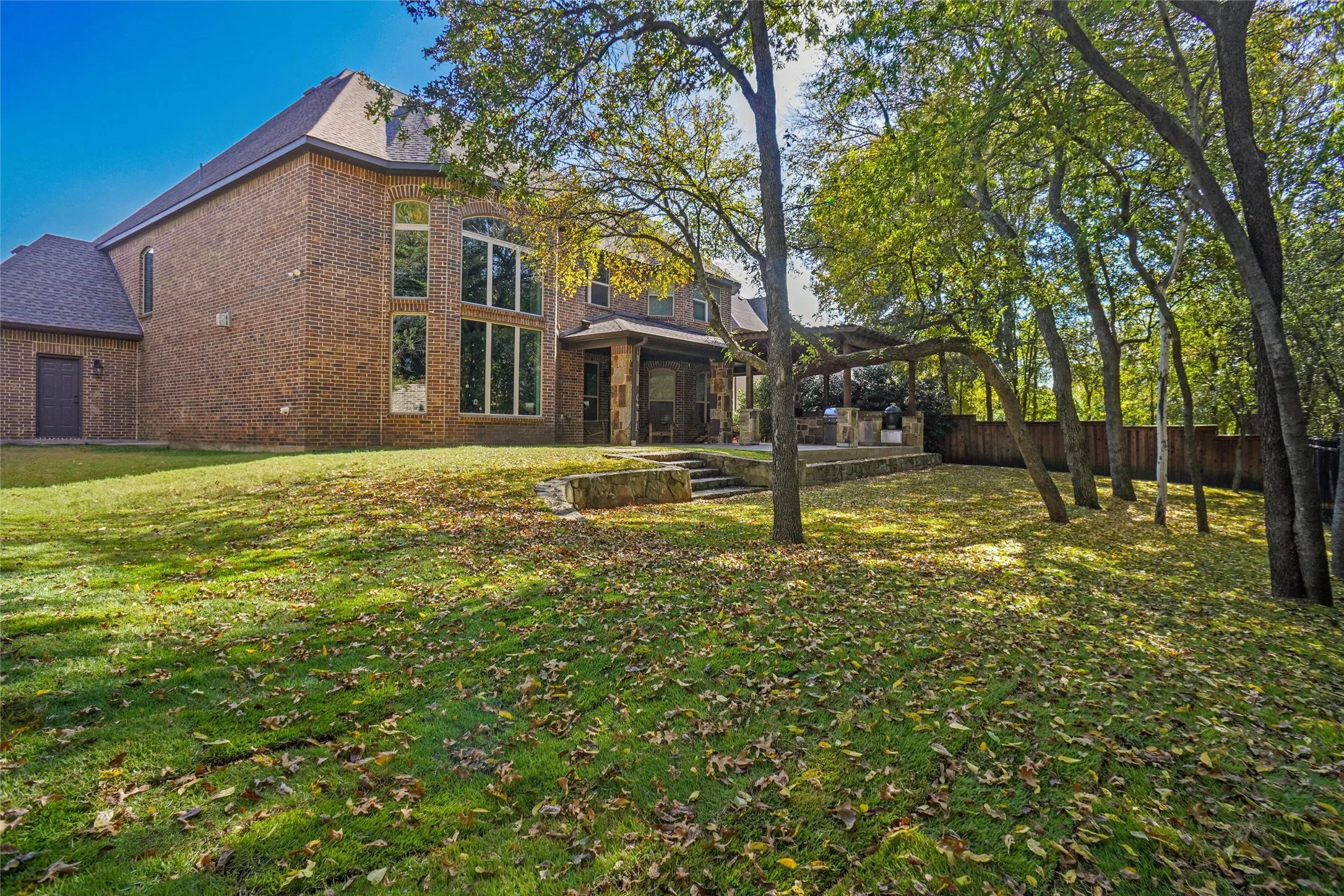 Brick exterior residence featuring a tiered lawn, mature trees, and a covered patio with a built-in outdoor kitchen