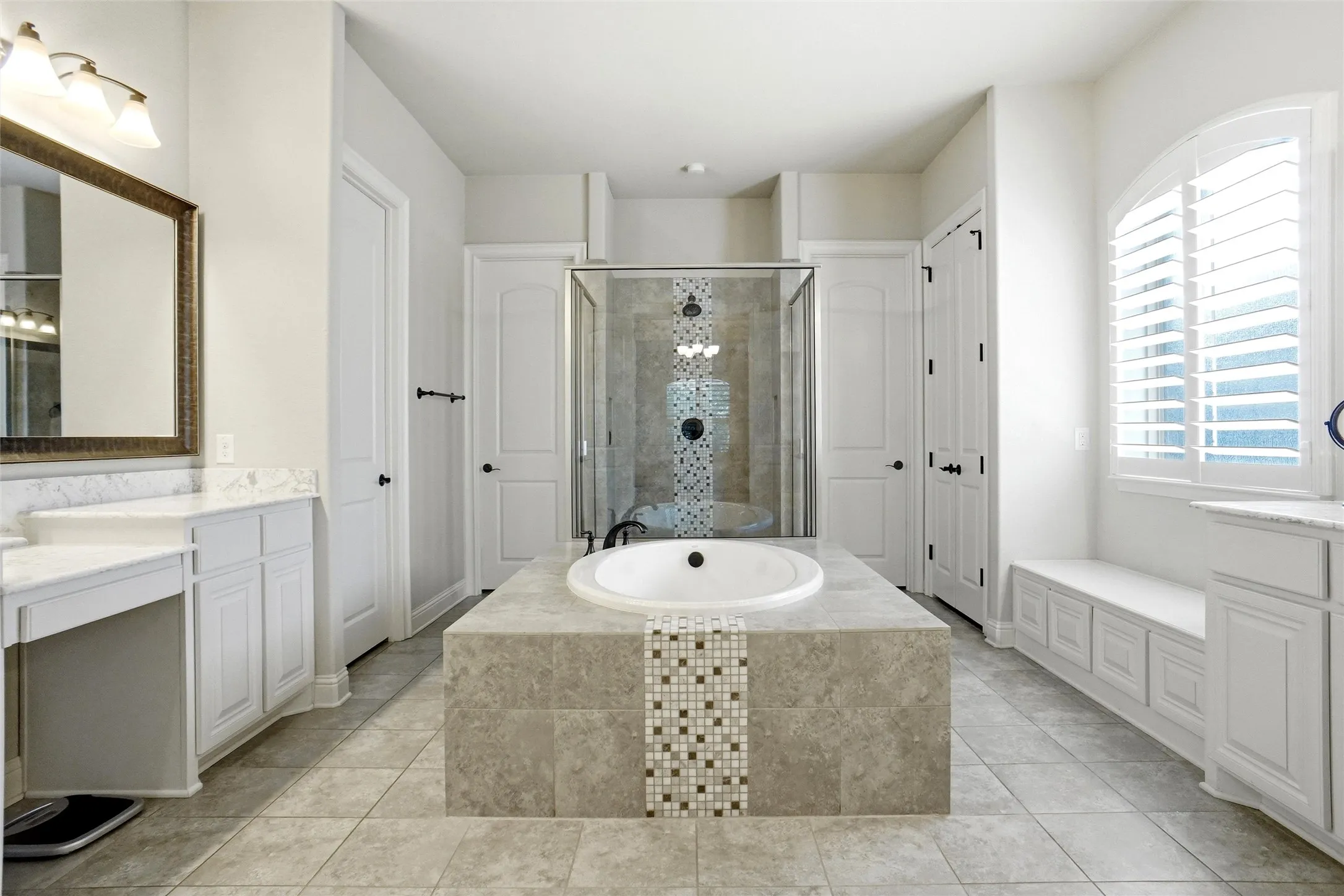 Spacious bathroom featuring a central built-in soaking tub with decorative tile accents, a glass-enclosed shower, dual vanity areas with white cabinetry and light-toned countertops, and a window with white louvered shutters