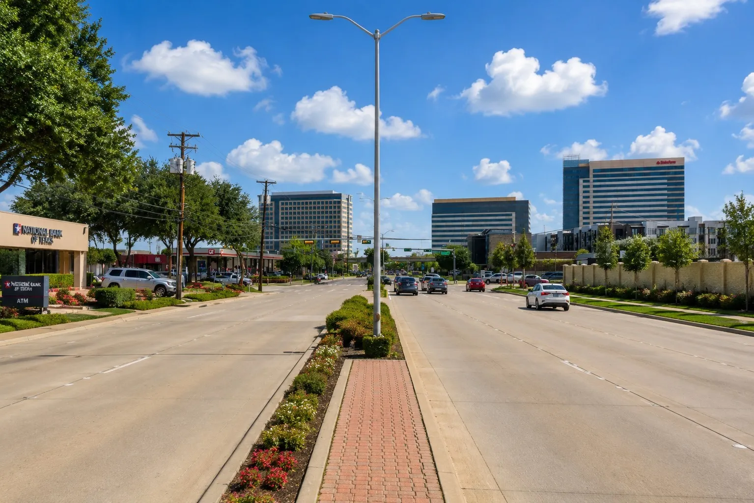 Commercial district featuring a multi-lane roadway, brick-paved median with landscaping, and a variety of retail and office buildings