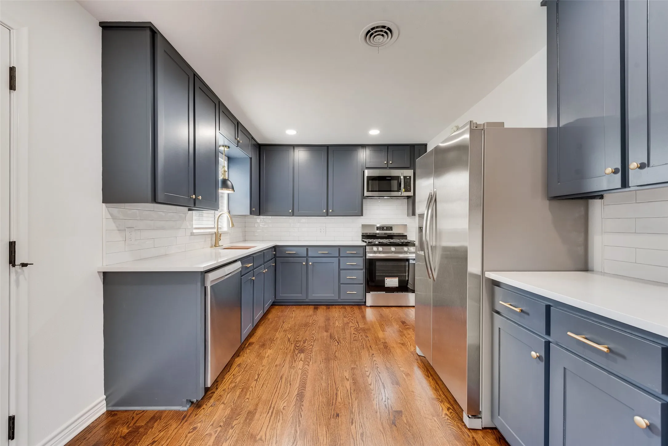 Kitchen featuring wood-finish flooring, white subway tile backsplash, light countertops, and dark cabinetry with brass hardware