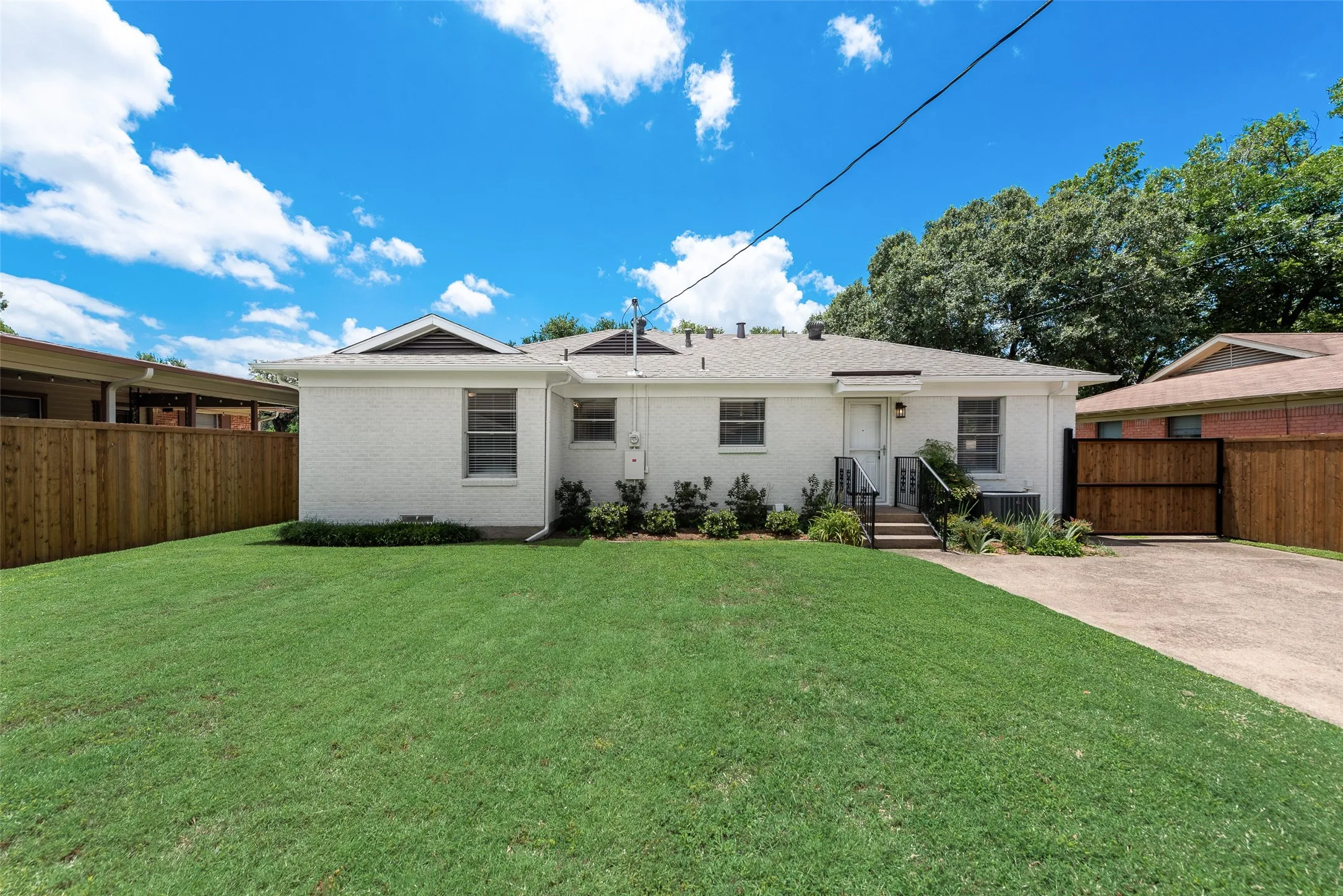 Expansive backyard featuring a lush lawn, white brick exterior, mature perimeter trees, wood privacy fencing, and a concrete driveway