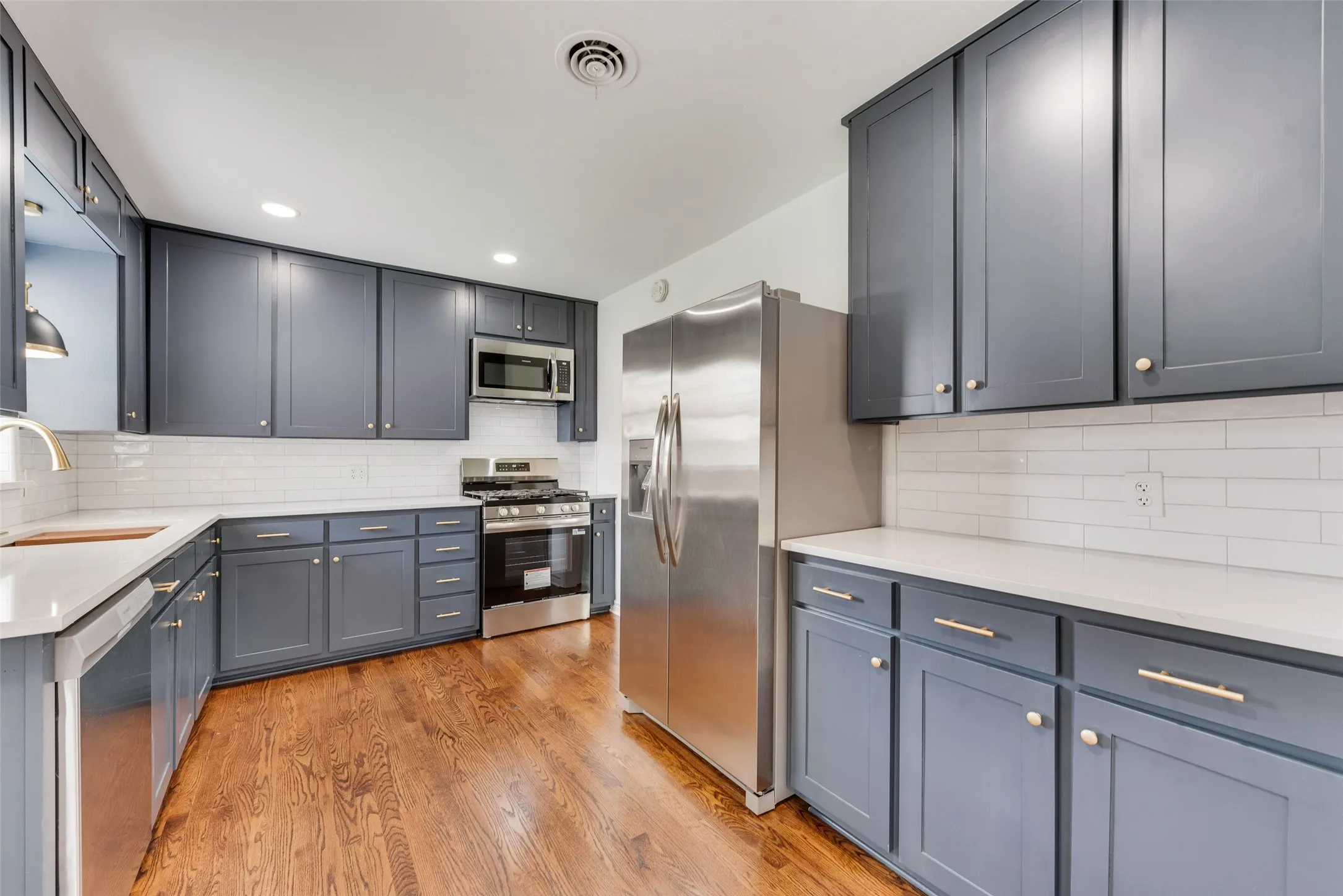 Kitchen featuring dark gray cabinetry, stainless steel appliances, a white subway tile backsplash, light-colored countertops, and wood-finish flooring