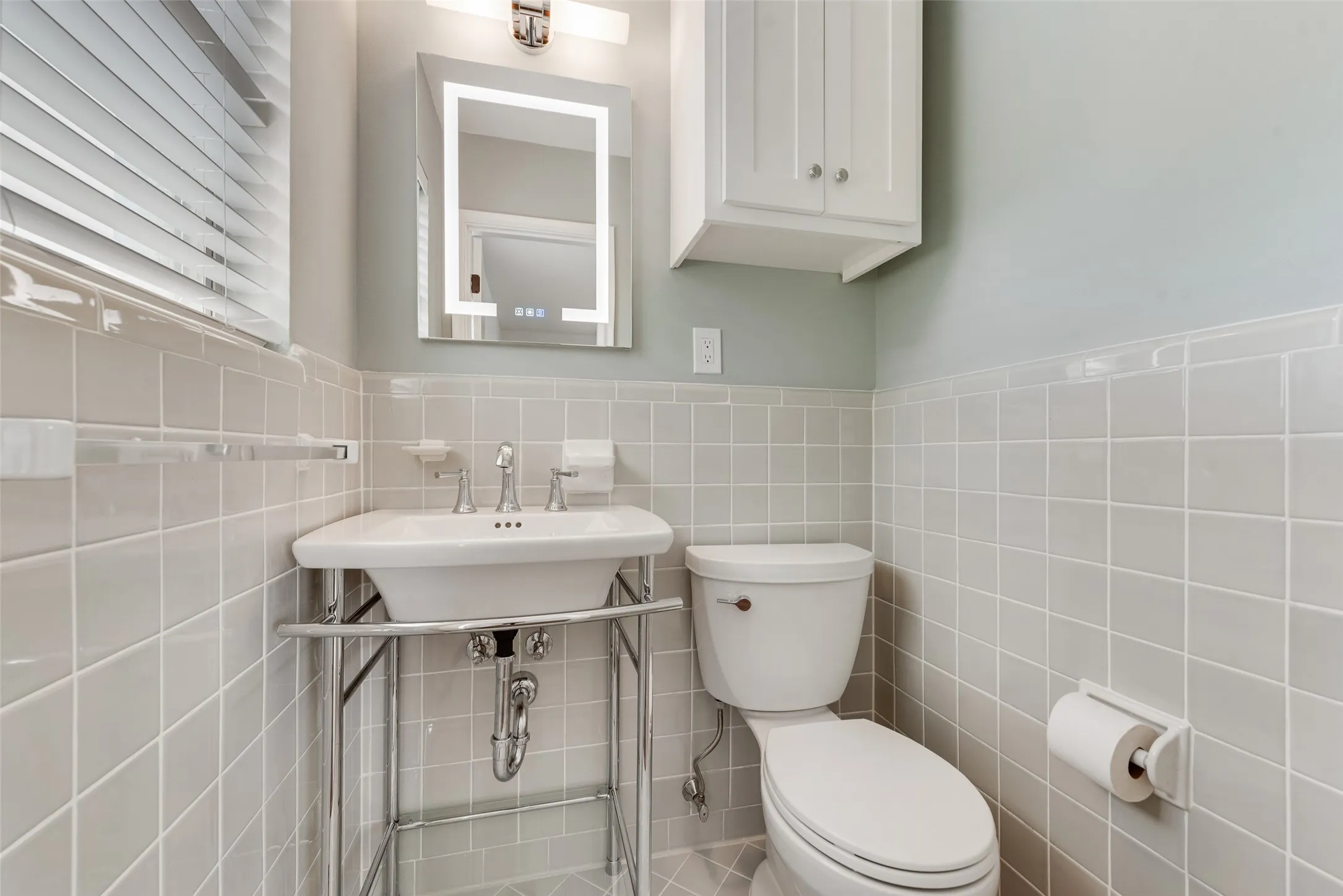 Bathroom featuring light grey square wall tiling, a pedestal sink with chrome stand, a wall-mounted lighted vanity mirror, and a white storage cabinet