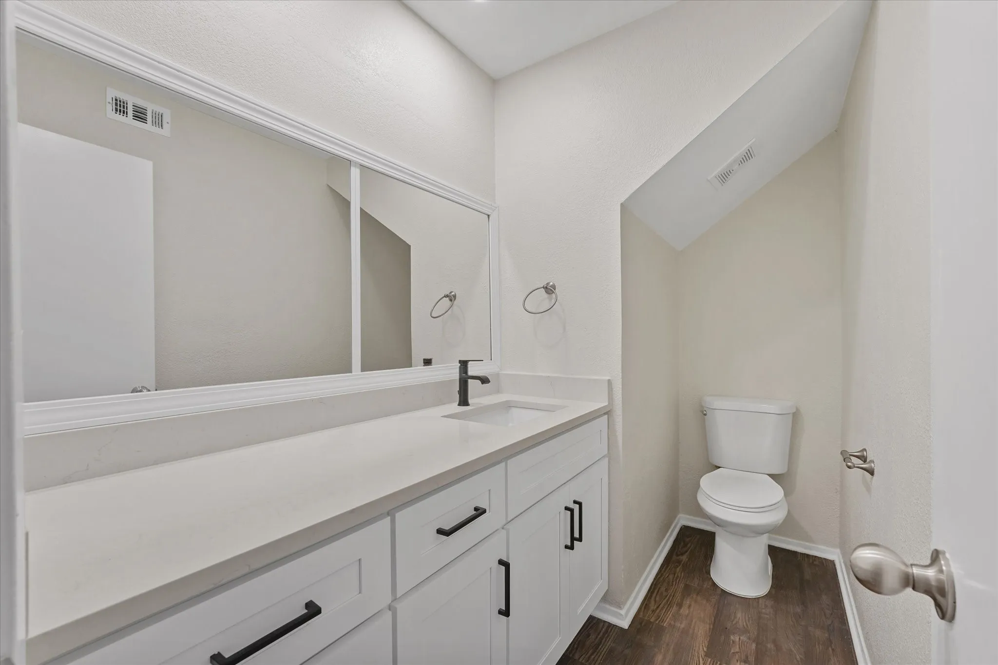 Bathroom featuring a white vanity with black hardware, a large wall-mounted mirror with decorative trim, a white countertop, a black single-handle faucet, and wood-finish flooring