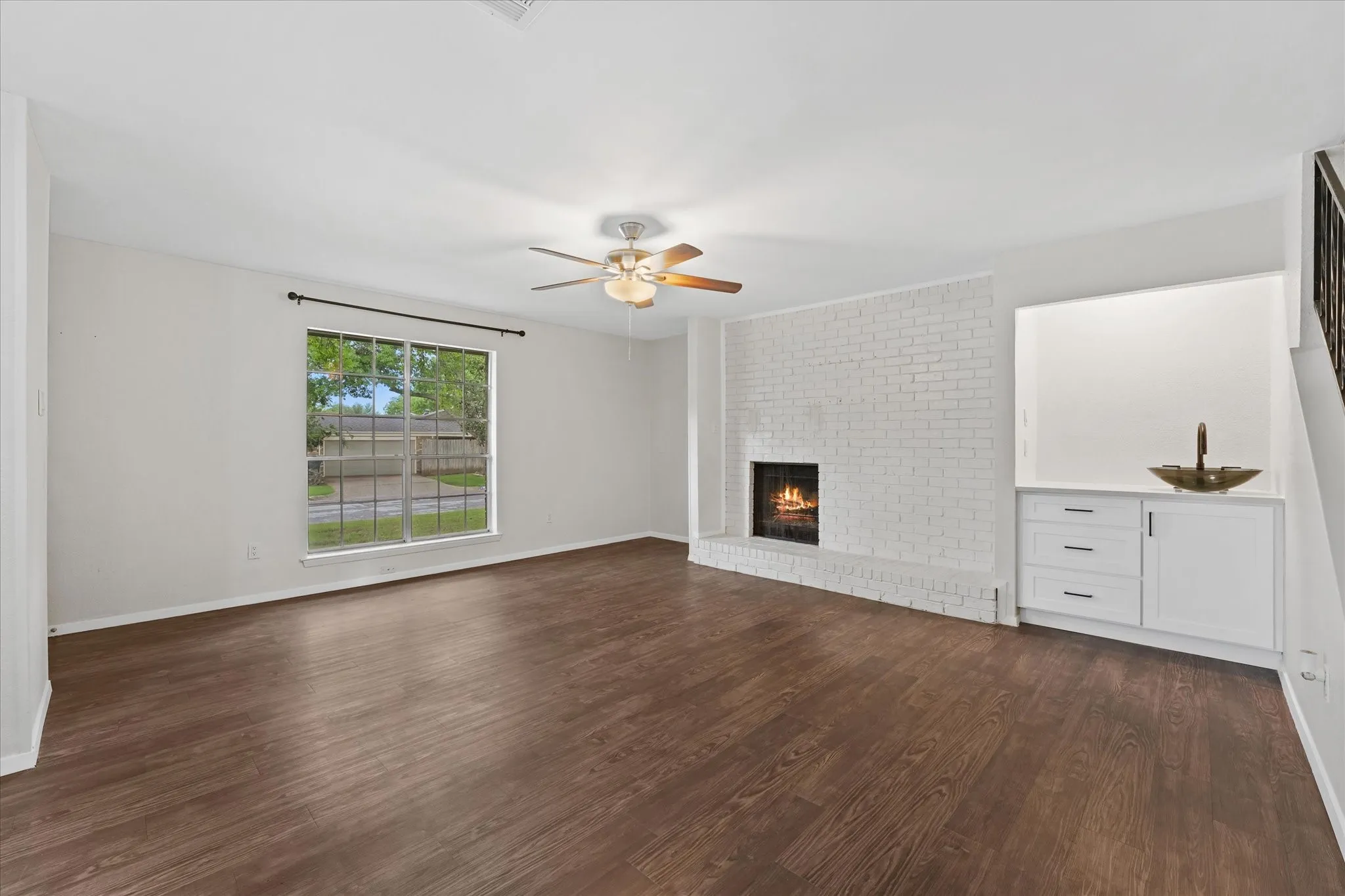 Spacious living area featuring dark wood-finish flooring, a white painted brick fireplace, and a built-in cabinetry unit with a white countertop