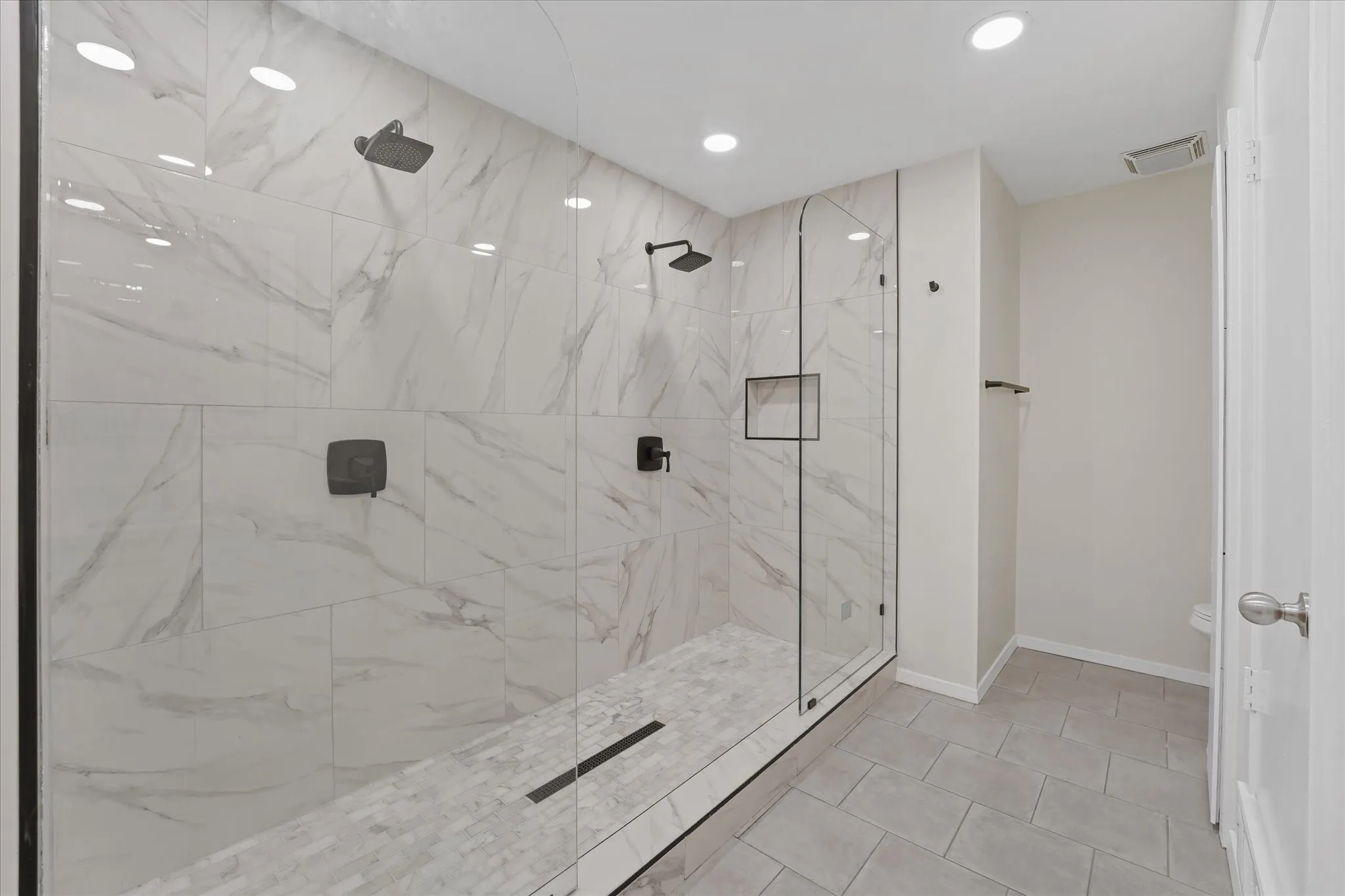 Spacious walk-in shower featuring clear glass enclosure, large format white porcelain wall tiles with gray veining, dual matte black rain shower heads, a recessed shower niche, and mosaic tile shower pan with linear drain