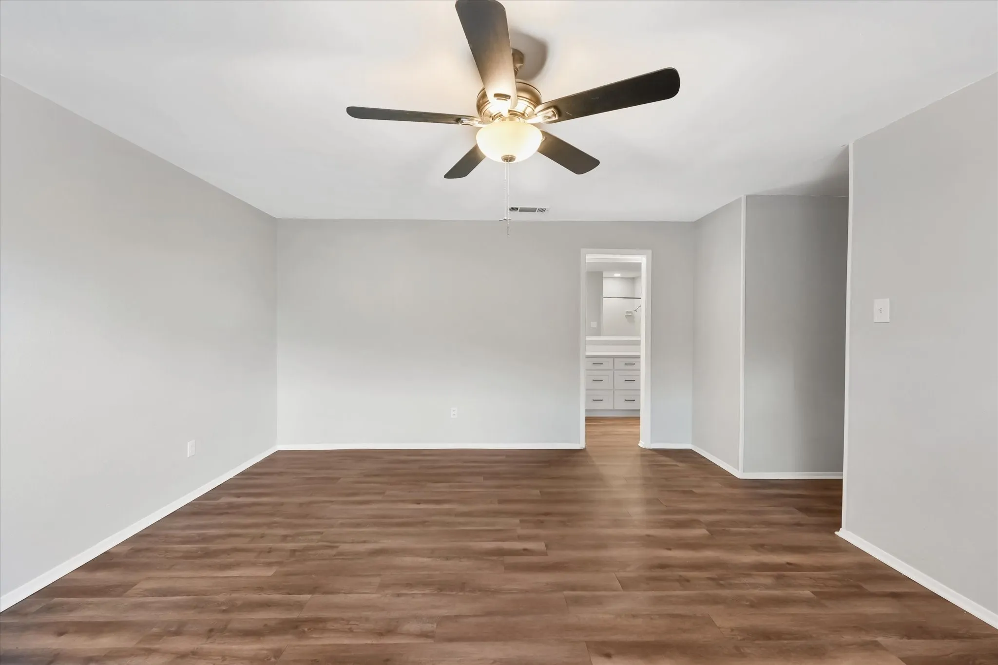 Spacious primary bedroom featuring wood-finish flooring, light grey painted walls, white baseboards, and a ceiling fan with integrated lighting