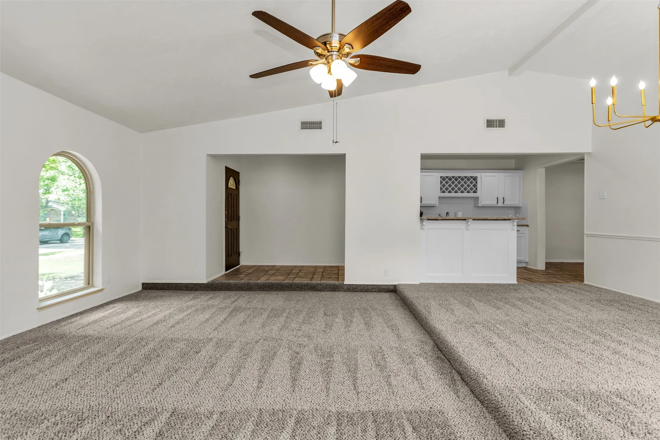 Spacious interior featuring a vaulted ceiling, light gray carpeting, an arched window, a ceiling fan with integrated lighting, and a gold-tone chandelier