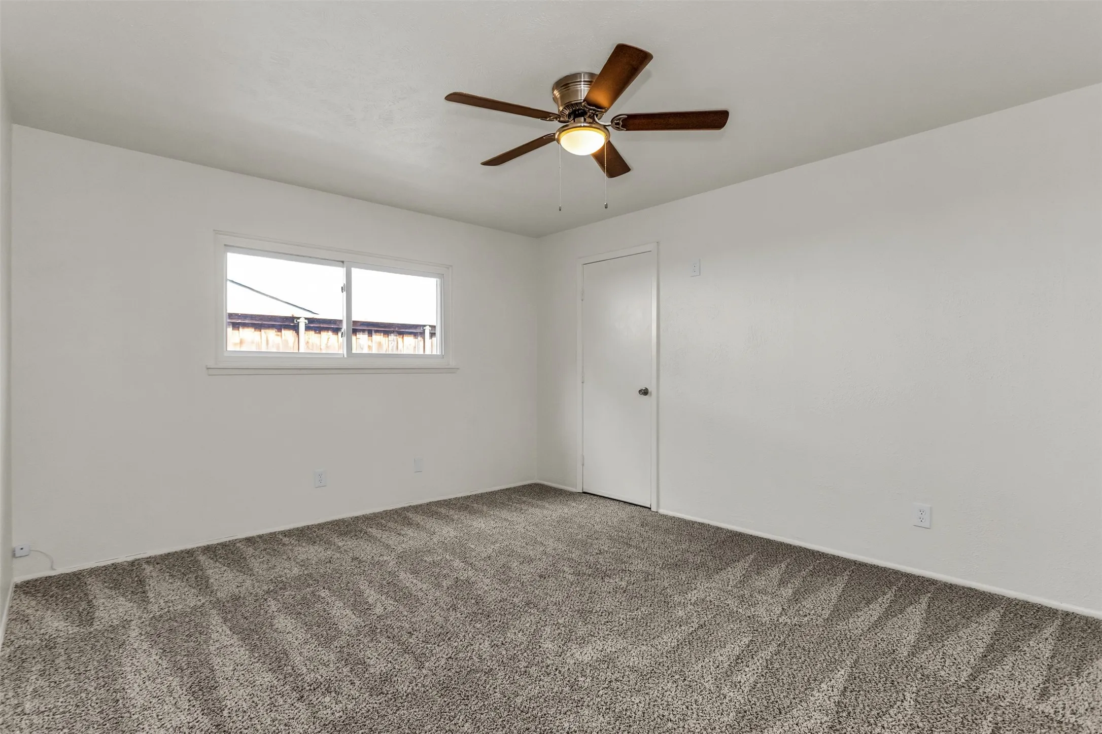 Spacious room featuring neutral carpeting, a dual-pane window with white trim, a ceiling fan with integrated lighting, and a white interior door