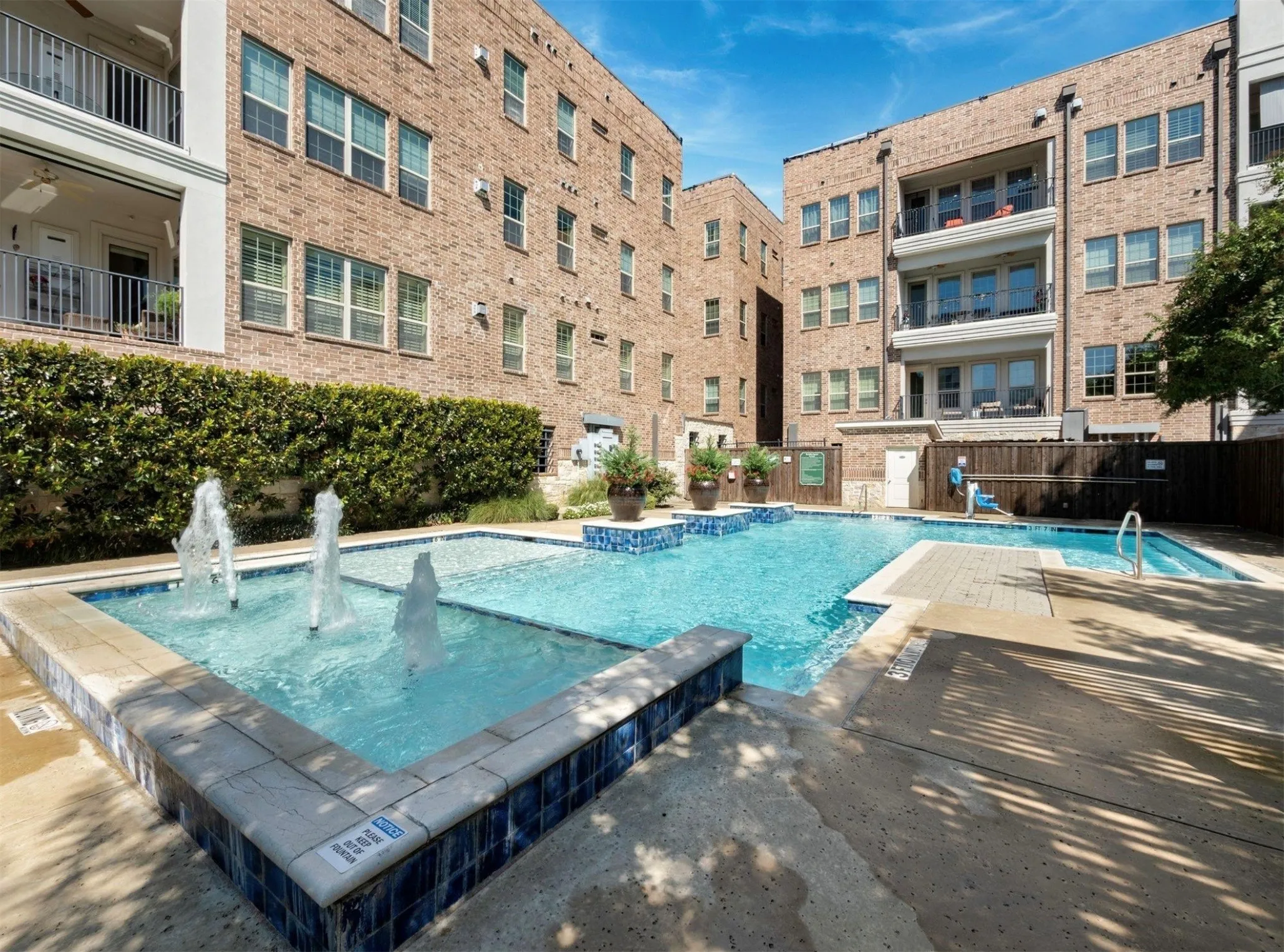 Outdoor swimming pool with integrated fountains, blue mosaic tile accents, surrounding concrete decking, and lush perimeter landscaping