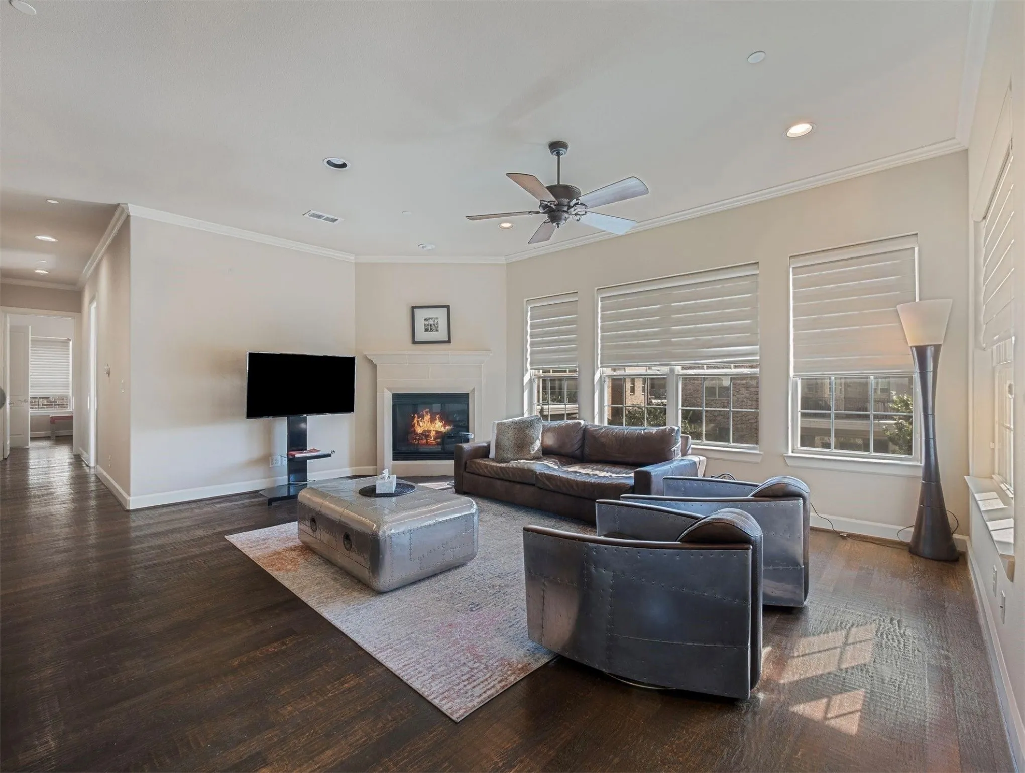 Spacious living area featuring dark wood-finish flooring, a corner fireplace with a mantel, multiple windows with blinds, crown molding, and recessed lighting