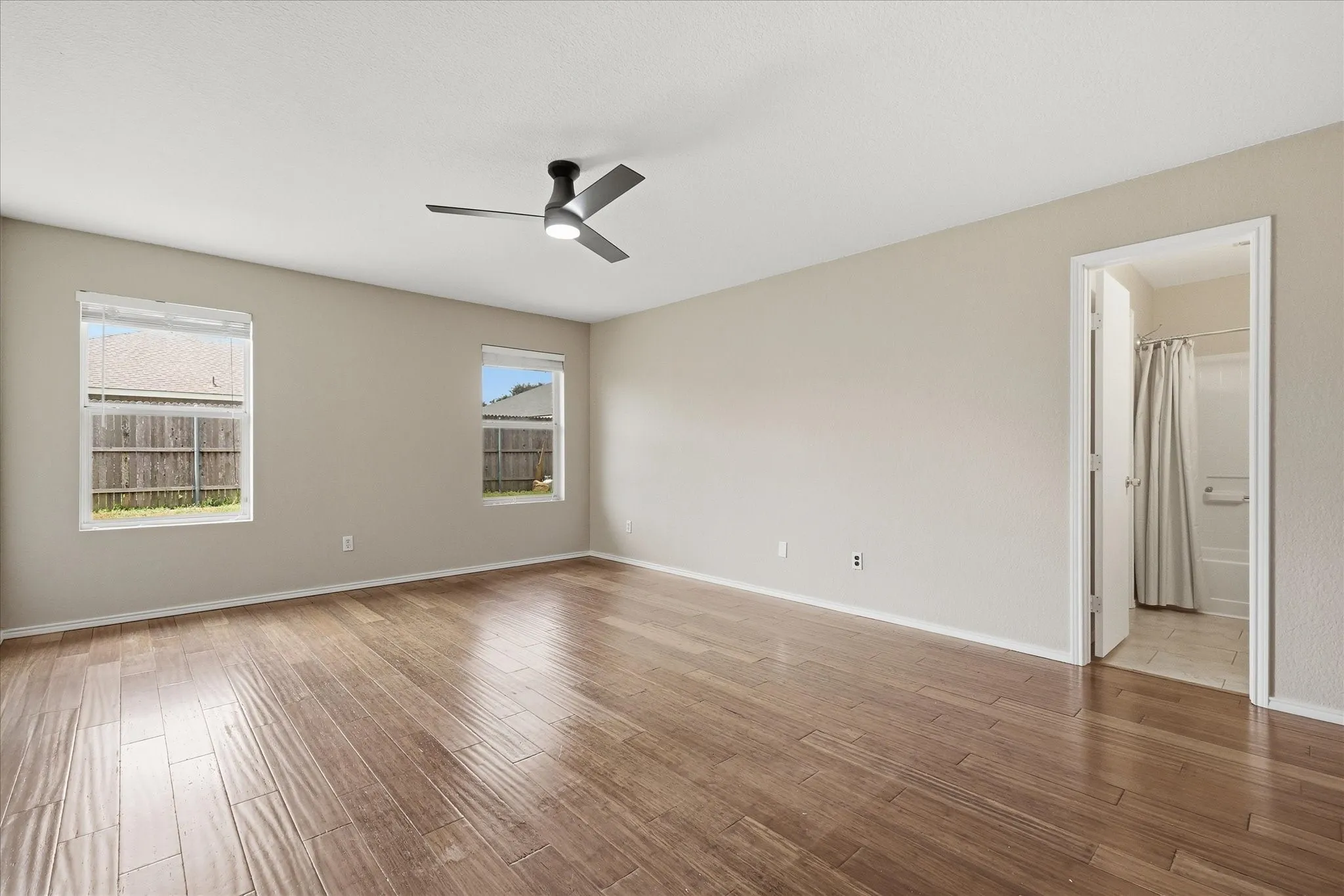 Spacious room featuring wood-finish flooring, two windows with blinds, a ceiling fan with integrated lighting, and neutral wall tones