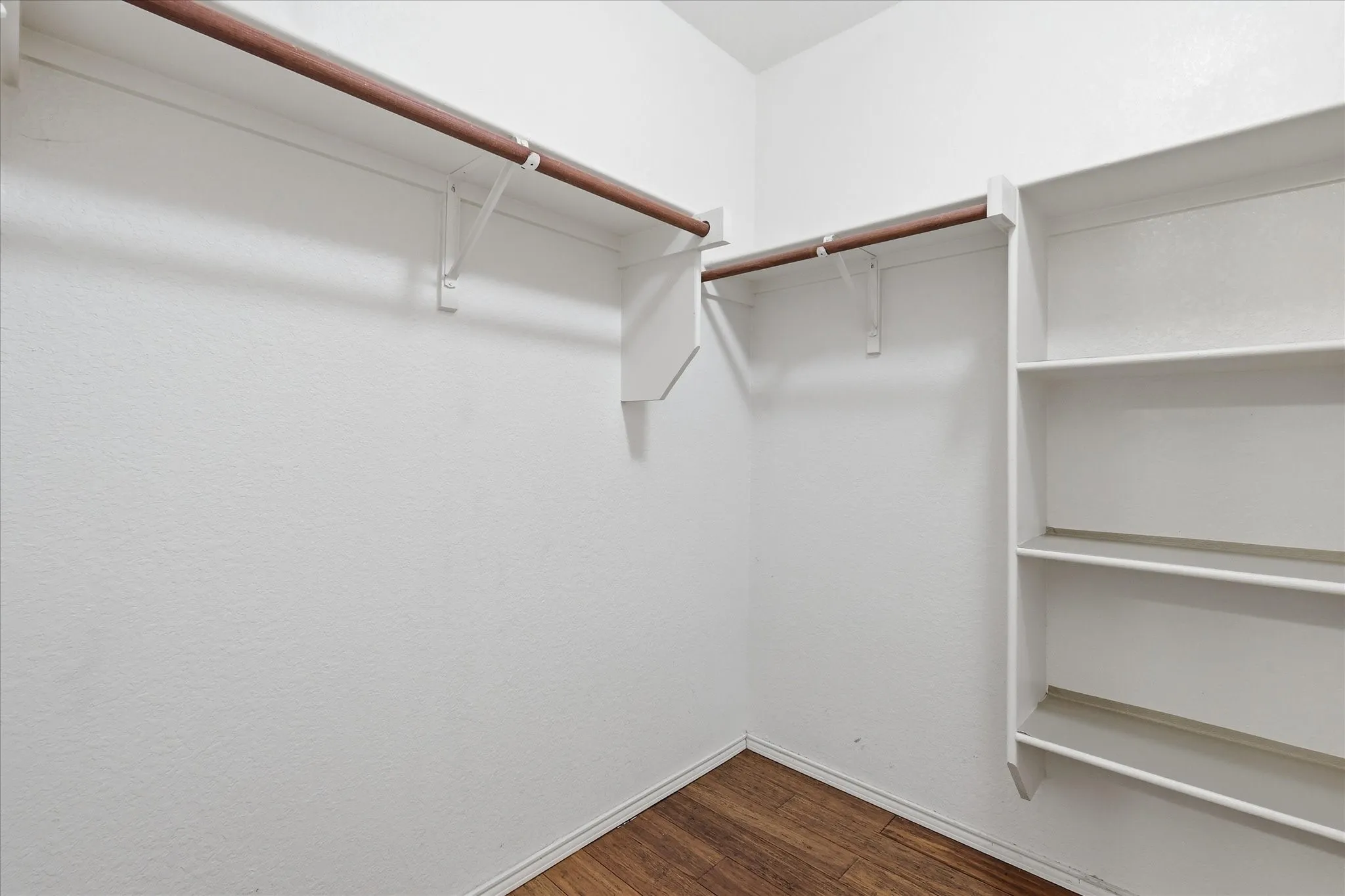 Spacious closet featuring wood-finish flooring, white painted walls, two wood-finish hanging rods, and built-in shelving with five shelves