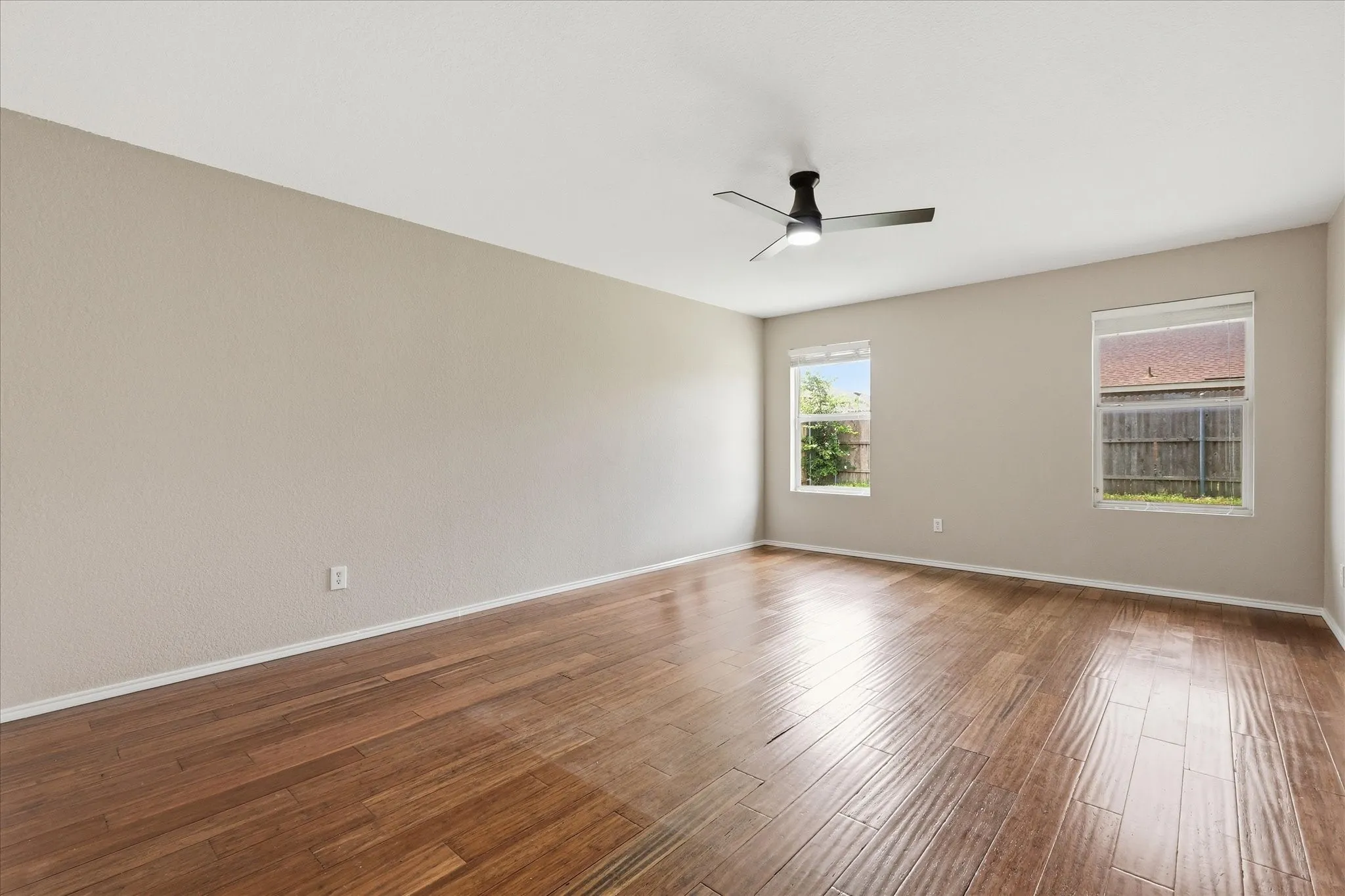 Spacious room featuring wood-finish flooring, two large windows, a ceiling fan with integrated lighting, textured walls, and white trim