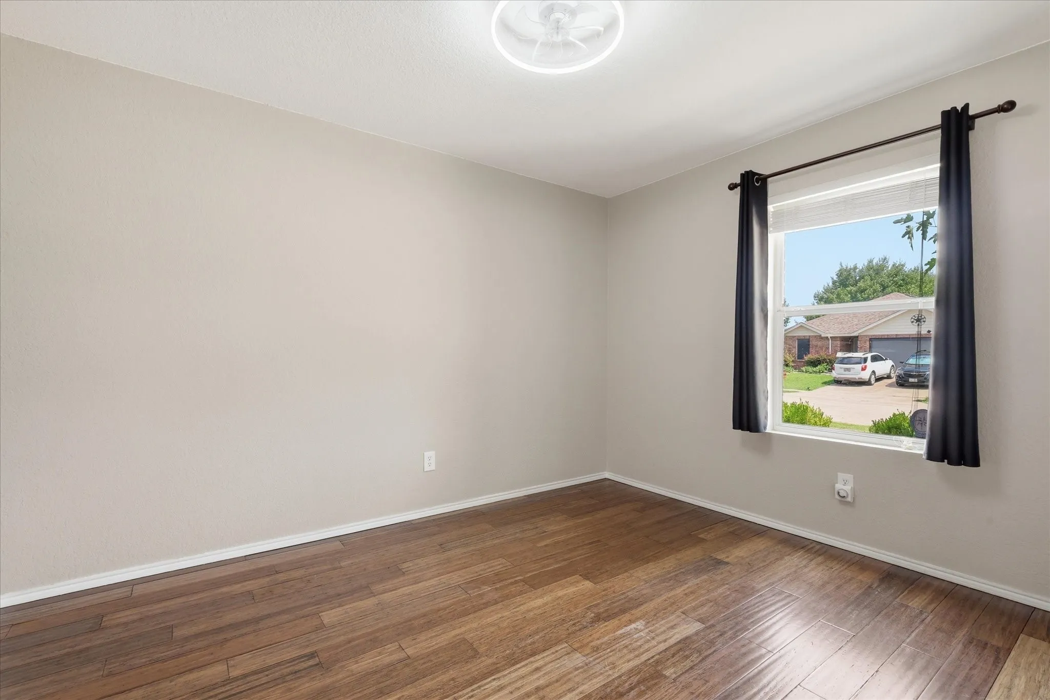 Spacious room featuring wood-finish flooring, neutral-toned walls, and a prominent window with dark curtain rods and drapes