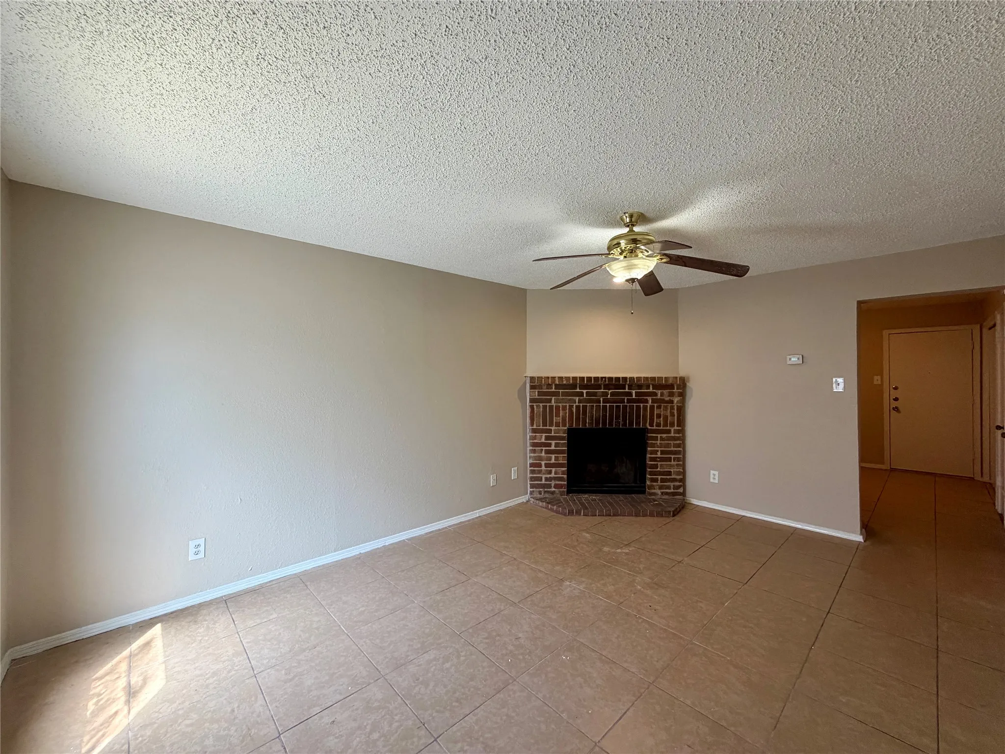 Tile flooring, corner brick fireplace, ceiling fan with light fixture, neutral wall tones, textured ceiling