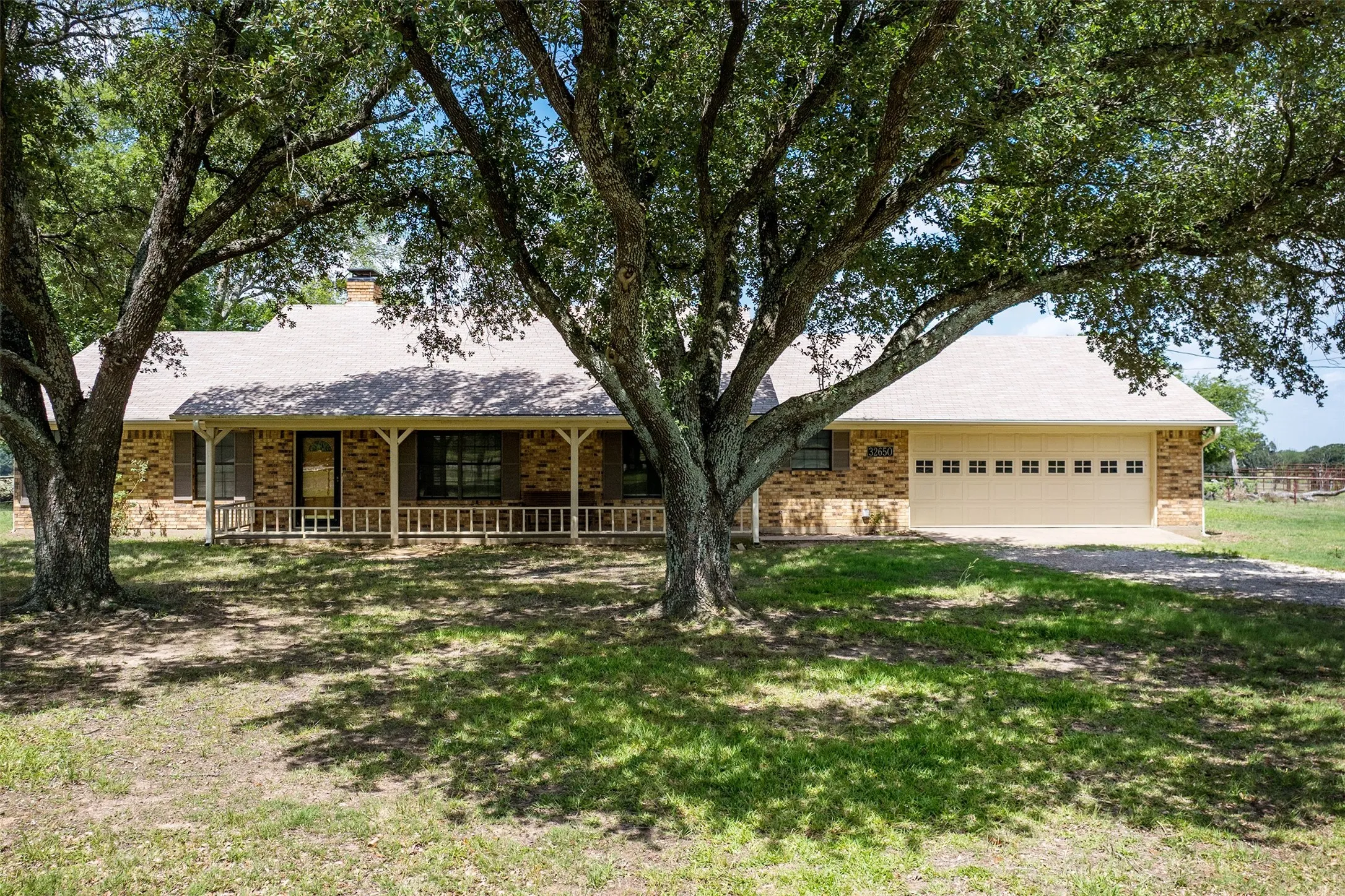 Brick exterior residence featuring a covered front porch with white railing, a light-colored shingled roof, a two-car garage with divided light windows, and mature trees providing natural shade