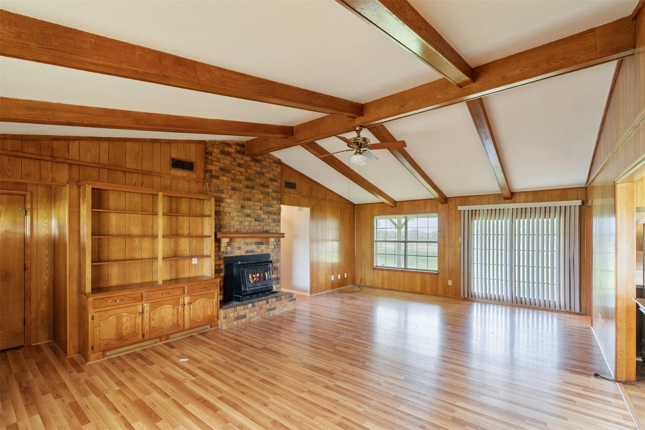 Spacious living area featuring wood-finish flooring, brick fireplace with wood stove insert, vaulted ceiling with exposed wood beams, built-in cabinetry, and sliding glass door