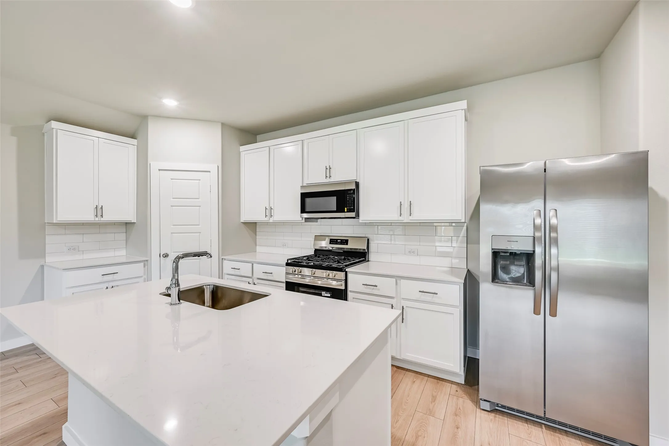 Kitchen featuring white shaker-style cabinetry, a large kitchen island with an undermount sink and gooseneck faucet, light-toned countertops, a stainless steel side-by-side refrigerator, and wood-finish flooring