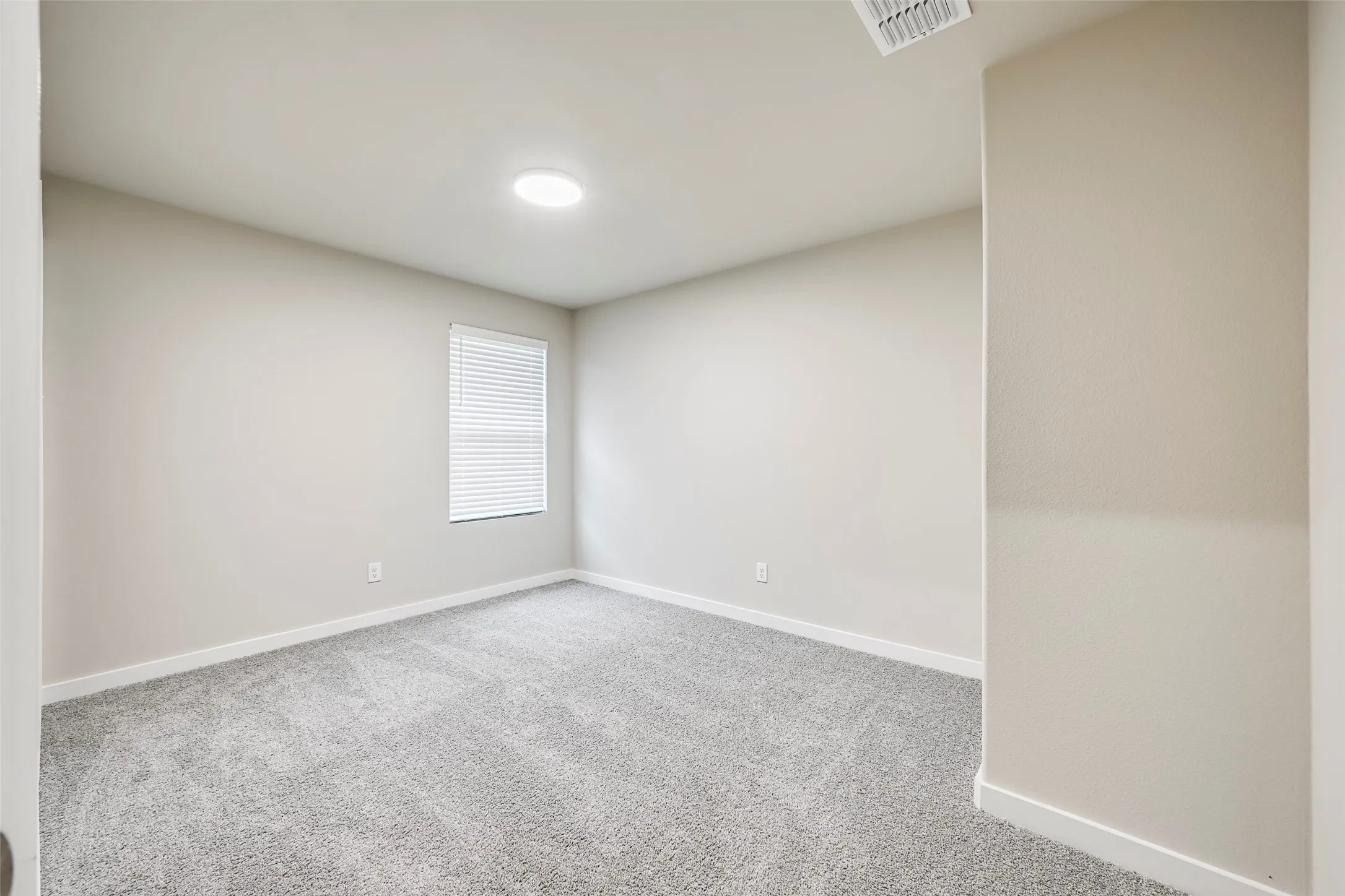 Neutral-toned room with light gray walls, gray carpet, a single window with blinds, and a circular ceiling light fixture