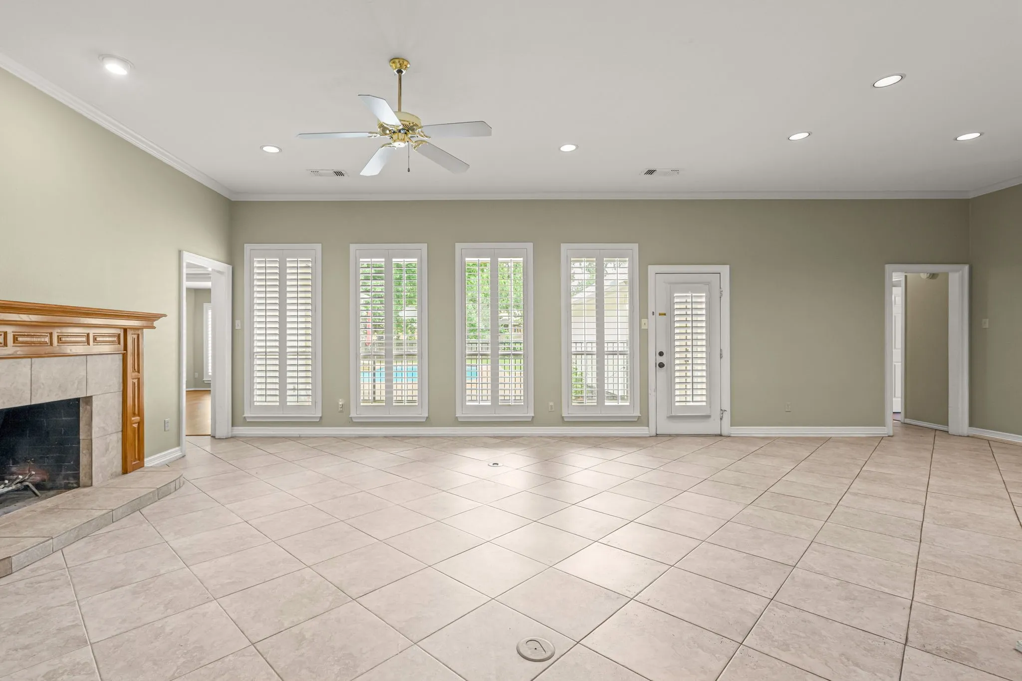 Spacious room featuring tile flooring, a fireplace with a wood mantel, multiple windows with white plantation shutters, a ceiling fan, and recessed lighting