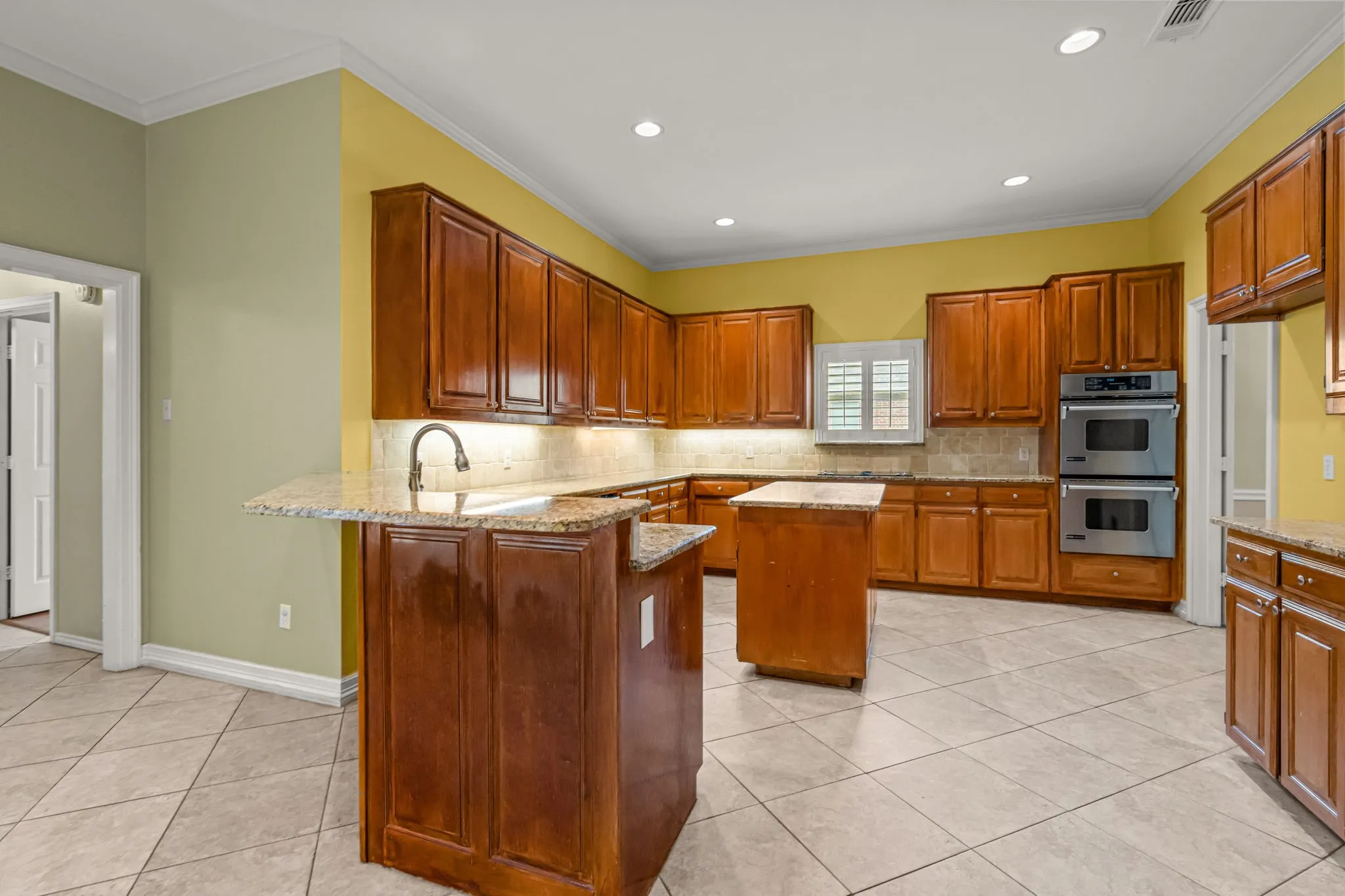 Spacious kitchen featuring warm wood cabinetry, light granite countertops, a central island, stainless steel double ovens, and tile flooring