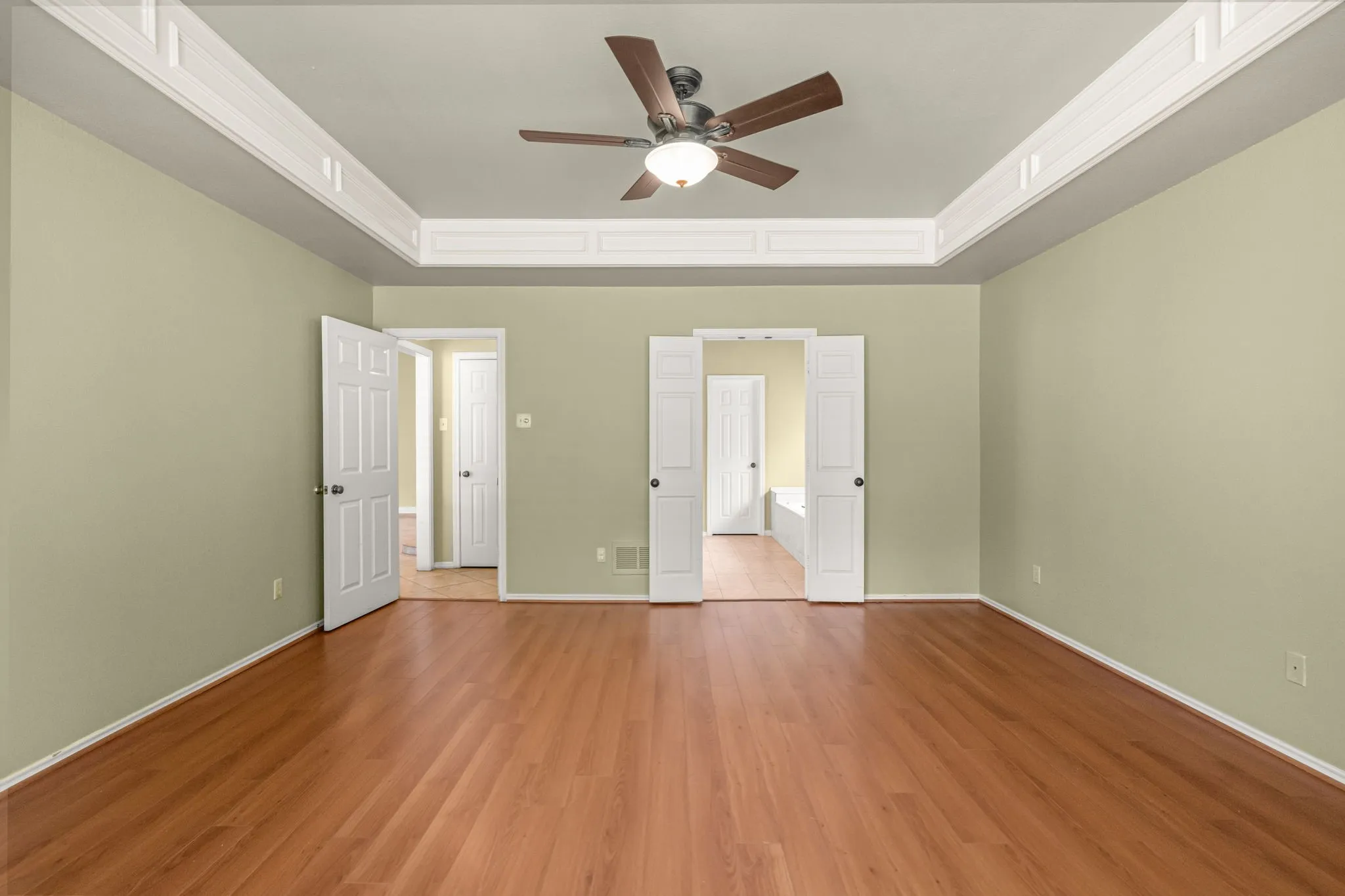 Spacious room featuring wood-finish flooring, a tray ceiling with decorative molding, and a ceiling fan with integrated lighting