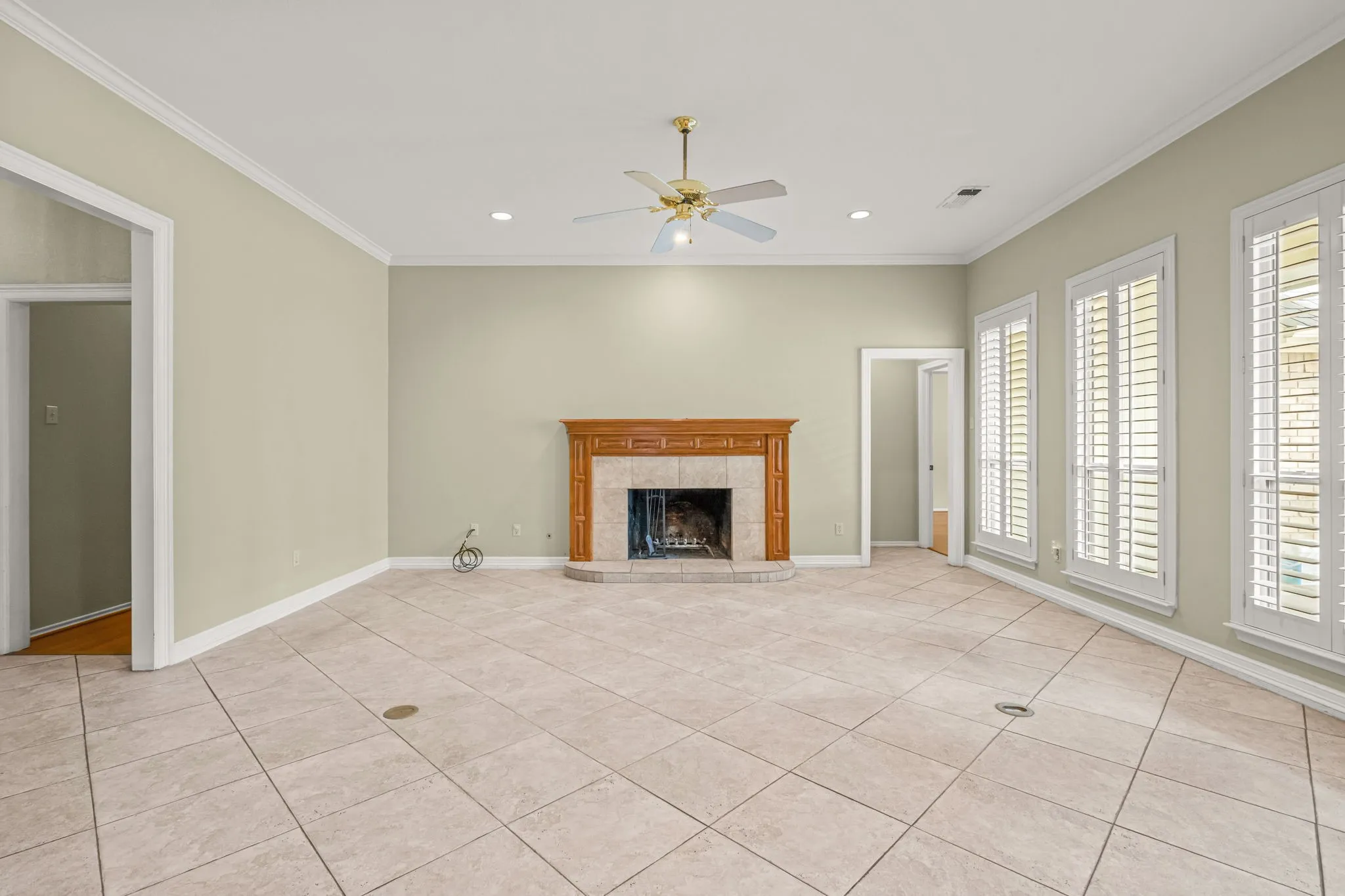 Spacious room featuring a central wood-mantle fireplace with a tiled hearth, light-toned tiled flooring, three tall windows with white plantation shutters, a ceiling fan with integrated lighting, and recessed lighting