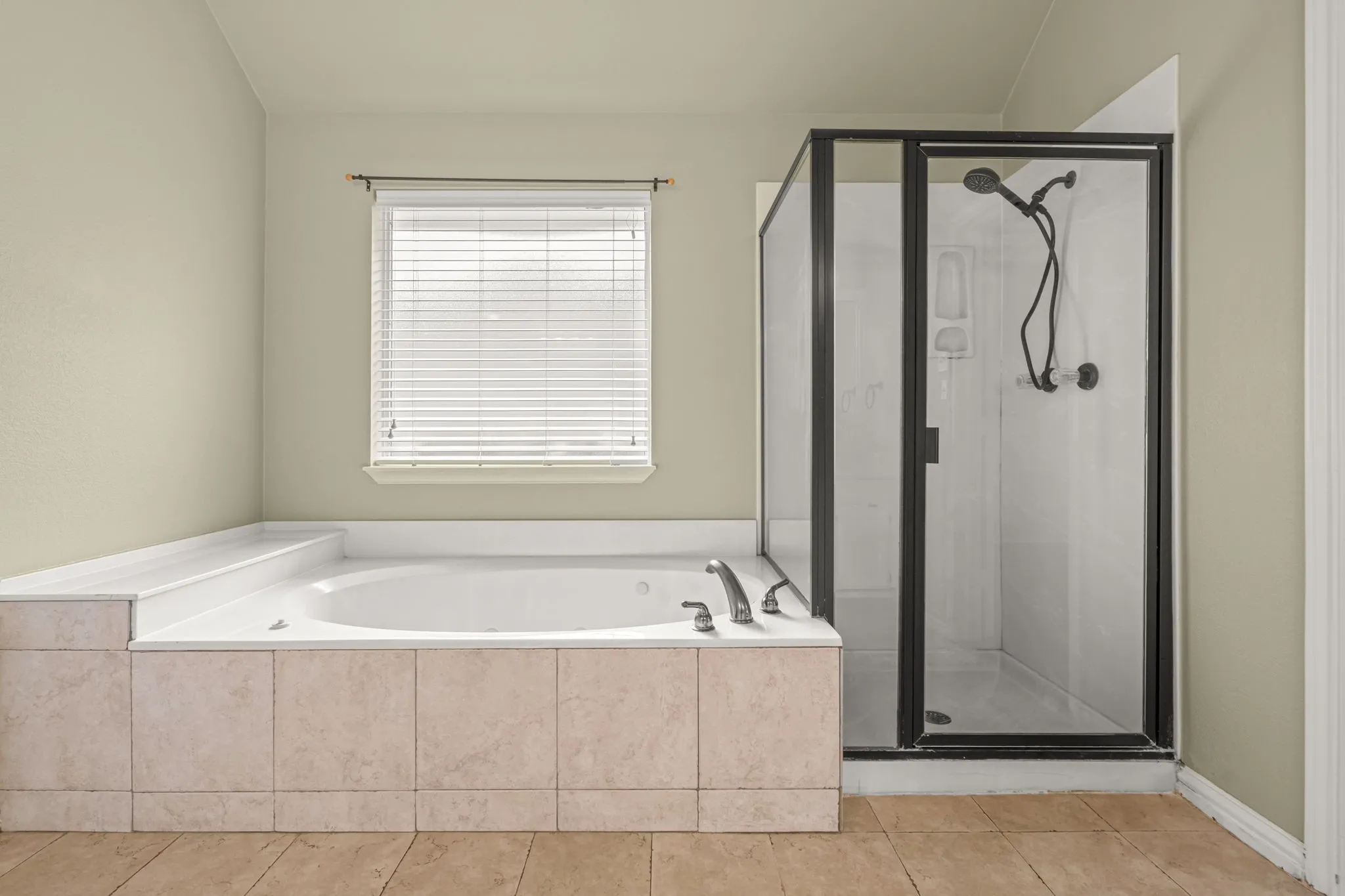 Soaking tub with tiled surround, separate enclosed shower with dark-framed glass, and window with blinds